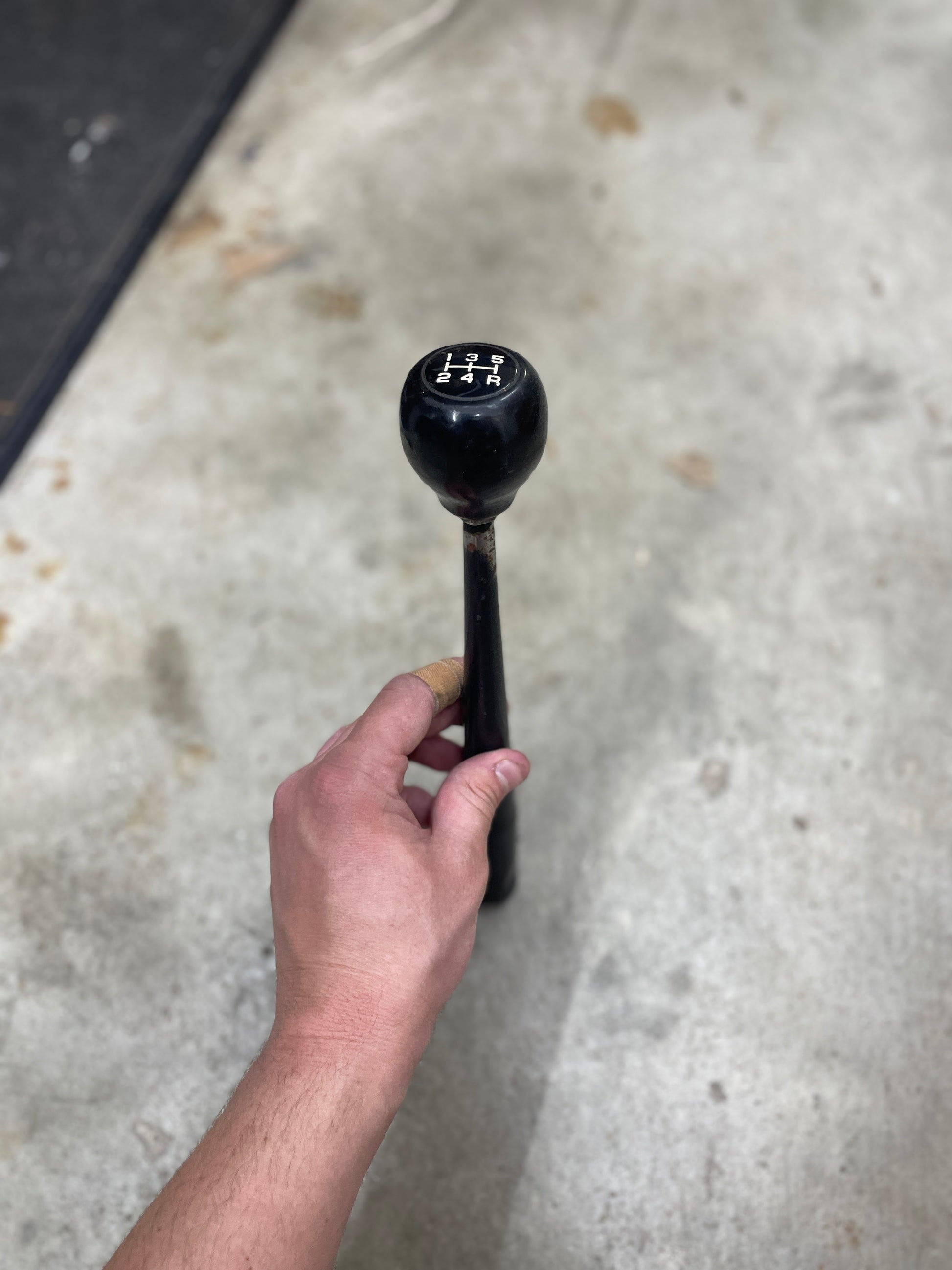 A hand holds a General Motors OEM 5 speed manual transmission shifter handle for 1982–1993 Chevy S10, Blazer, and more, with the shift pattern visible against a concrete floor background.
