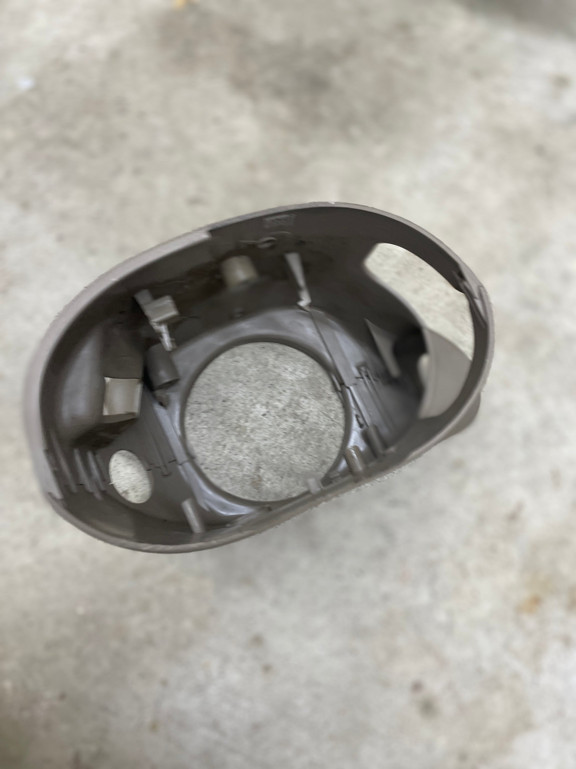 A close-up of a beige General Motors SET steering column shift cover for automatic transmissions (2001-2002 GMC Denali, Cadillac Escalade & more), oval-shaped with notches and an open center, held over concrete.
