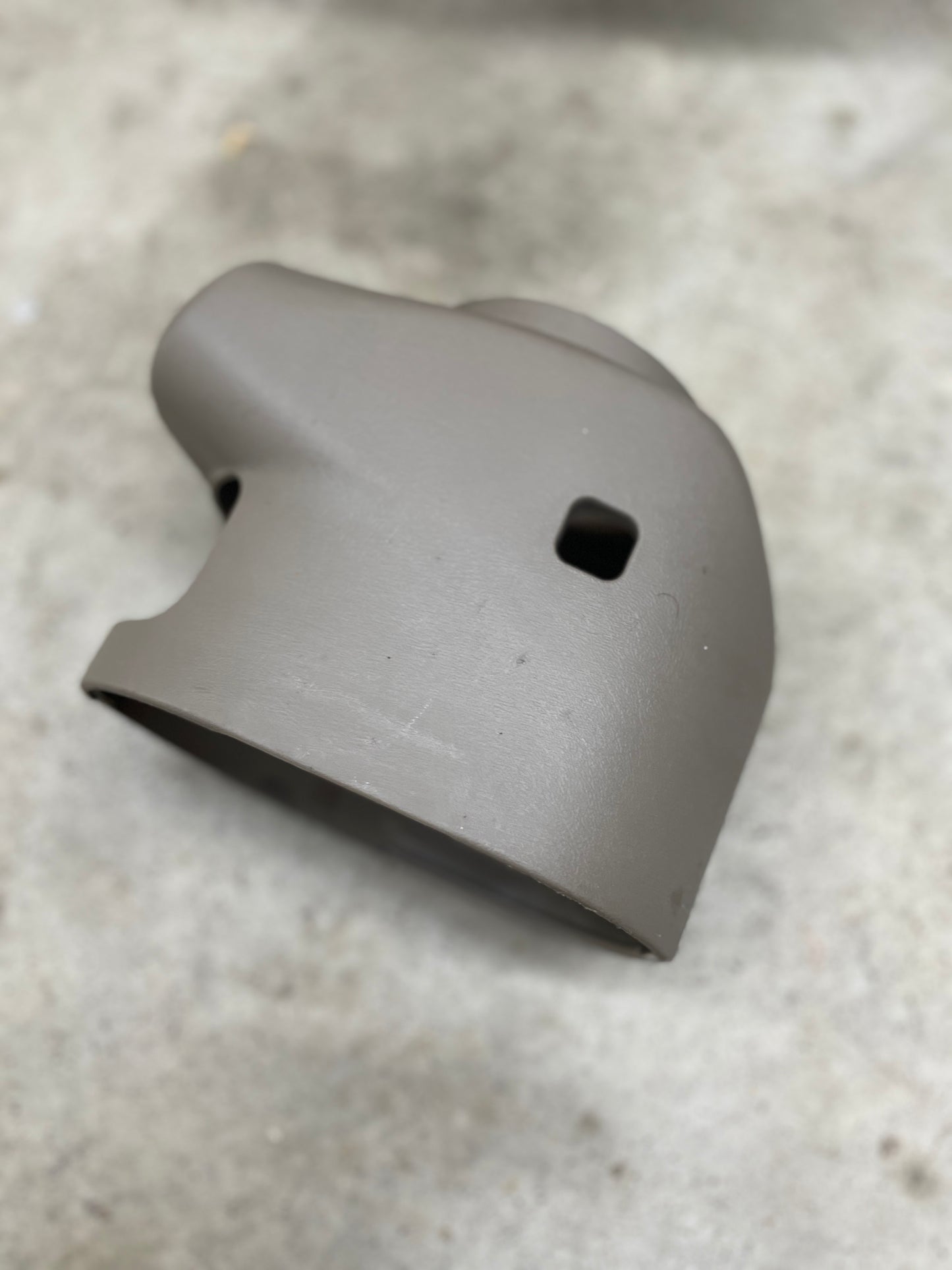 A beige SET Steering Column Shift Cover for automatic transmission by General Motors, designed for 2001-2002 GMC Denali, Cadillac Escalade, and more, rests on a light gray rough surface. It features two square holes and a cylindrical opening.