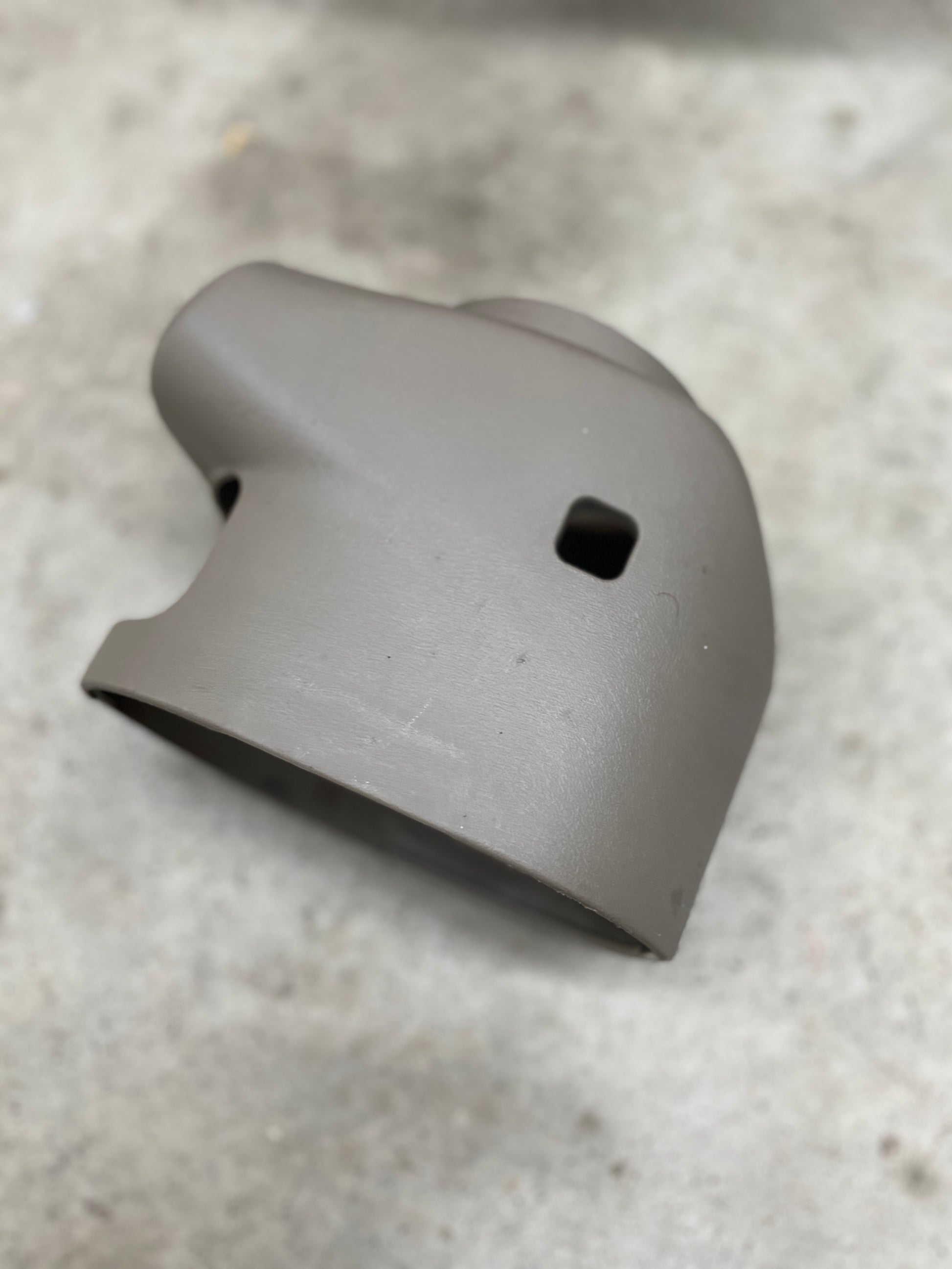 A beige SET Steering Column Shift Cover for automatic transmission by General Motors, designed for 2001-2002 GMC Denali, Cadillac Escalade, and more, rests on a light gray rough surface. It features two square holes and a cylindrical opening.