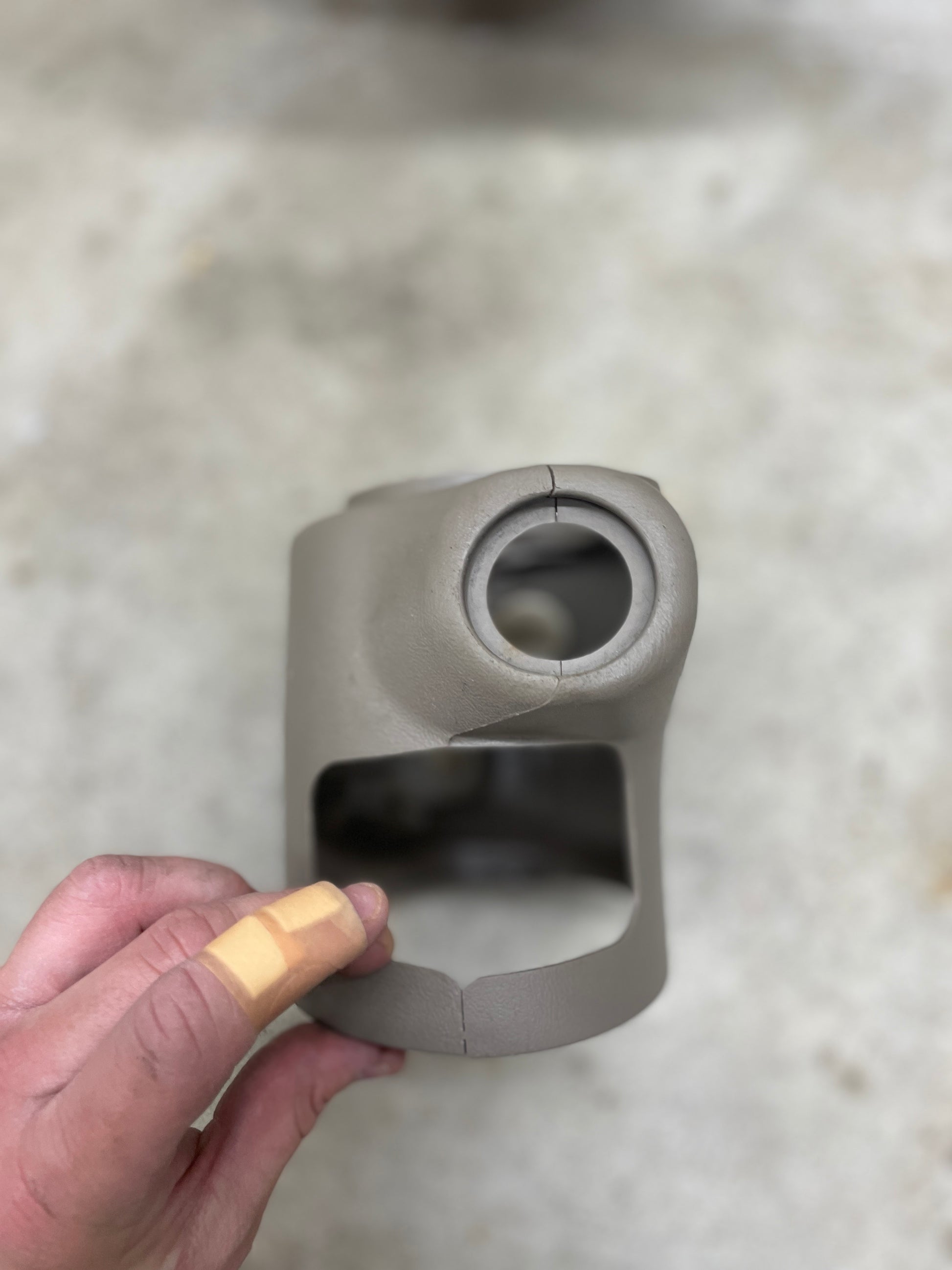 A bandaged hand holds a cracked General Motors SET Steering Column Shift Cover w/Automatic Transmission in Beige, for 2001-2002 GMC Denali and Cadillac Escalade, showing damage against a blurred light background.