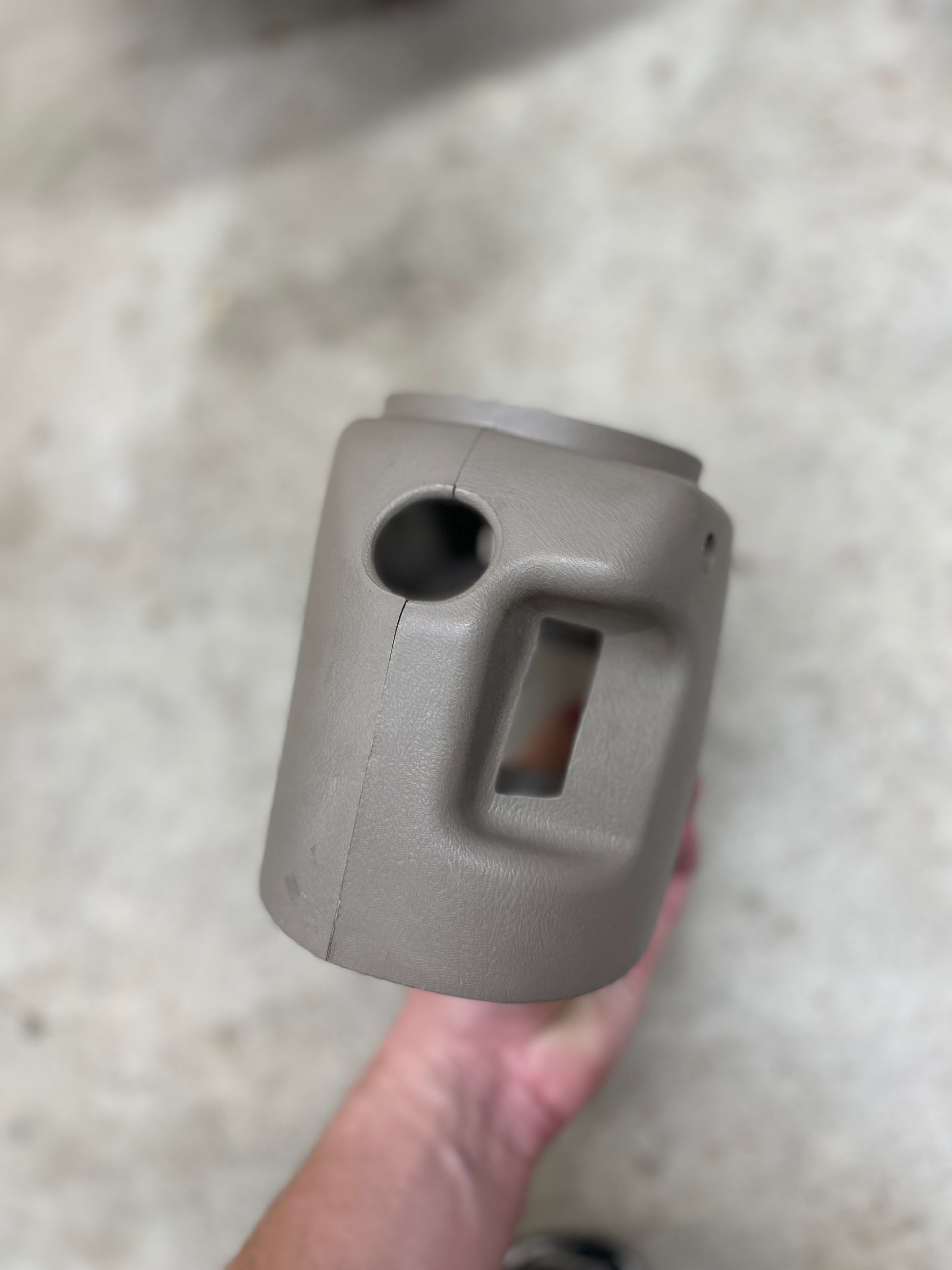 A hand holds a beige SET Steering Column Shift Cover w/Automatic Transmission by General Motors, featuring a handle and openings, perfect for 2001-2002 GMC Denali, Cadillac Escalade, and more—set against a blurred light background.