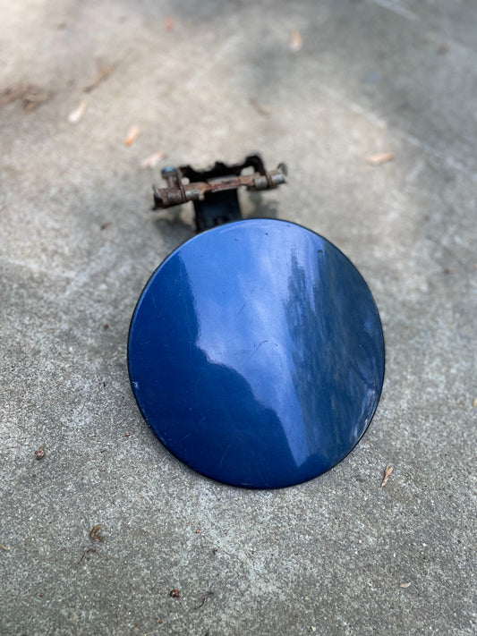 A General Motors OEM Fuel Filler Neck Door Gas Tank Cover in blue for 2001-2004 Chevy S10 & GMC Sonoma, featuring a rusty hinge, lies outdoors on concrete and reflects trees and sky.
