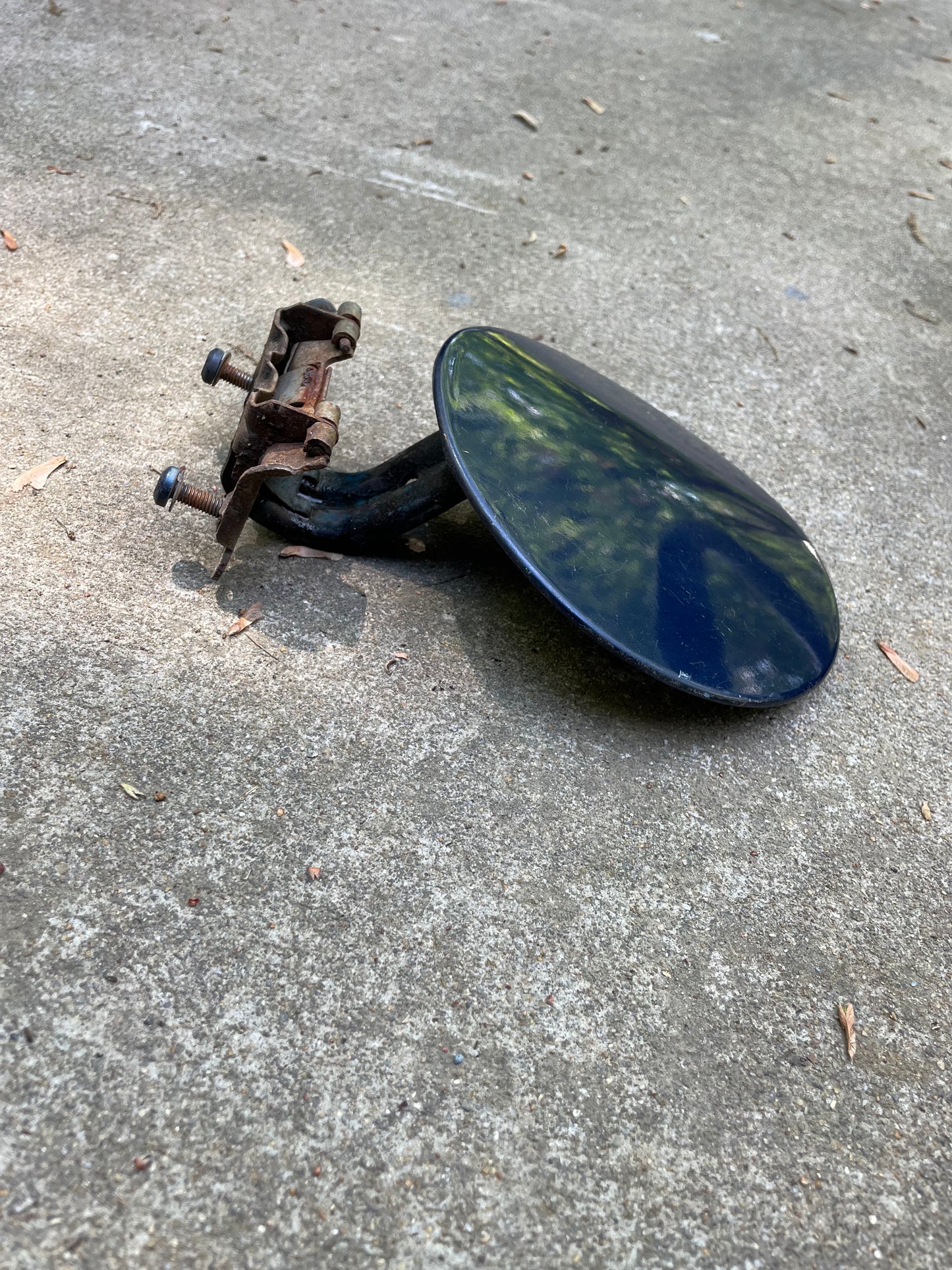 A round, black, metal side mirror with a rusty bracket rests on rough concrete next to a General Motors OEM Fuel Filler Neck Door Gas Tank Cover in blue for 2001-2004 Chevy S10 & GMC Sonoma.