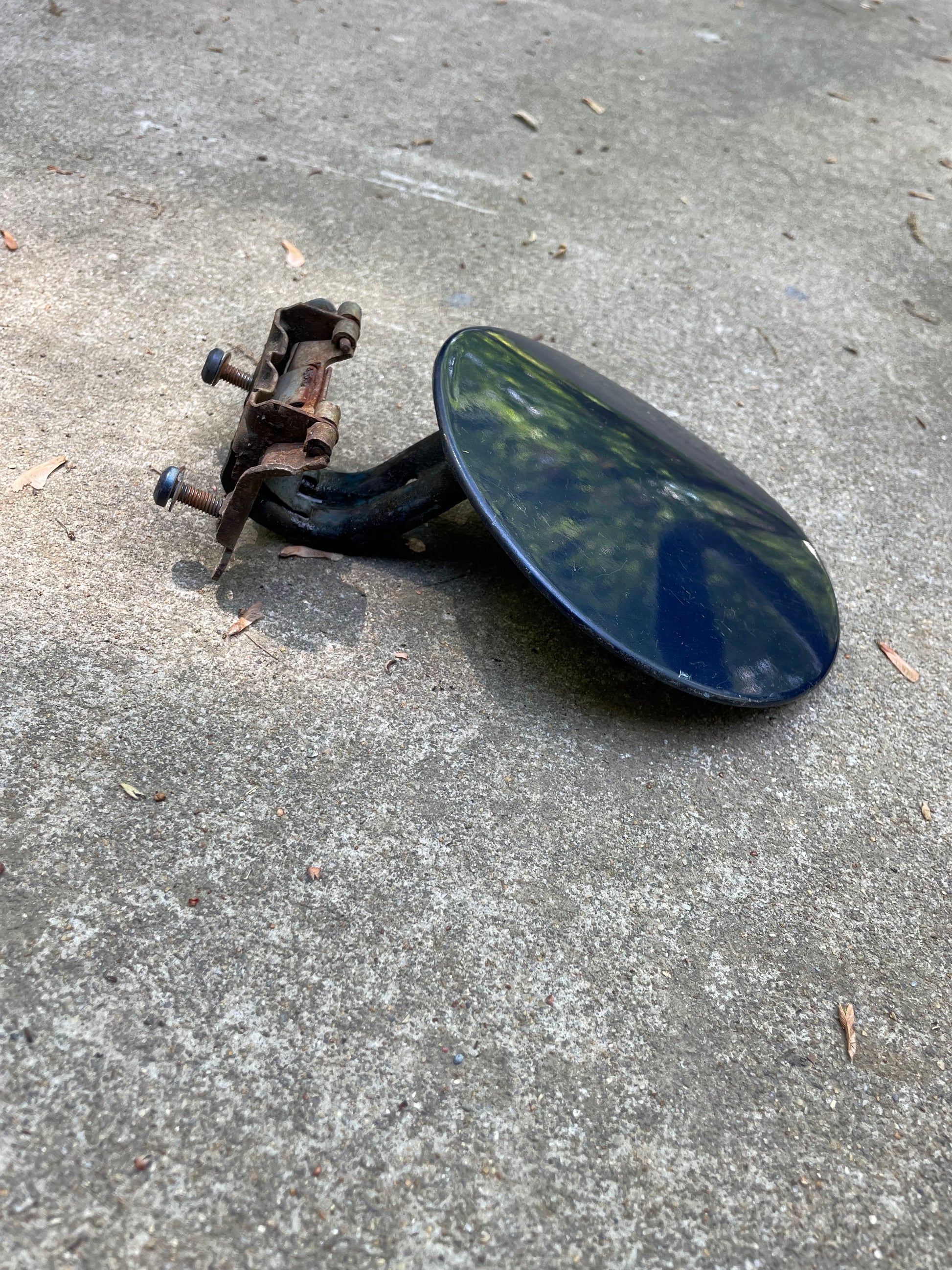 A round, black, metal side mirror with a rusty bracket rests on rough concrete next to a General Motors OEM Fuel Filler Neck Door Gas Tank Cover in blue for 2001-2004 Chevy S10 & GMC Sonoma.