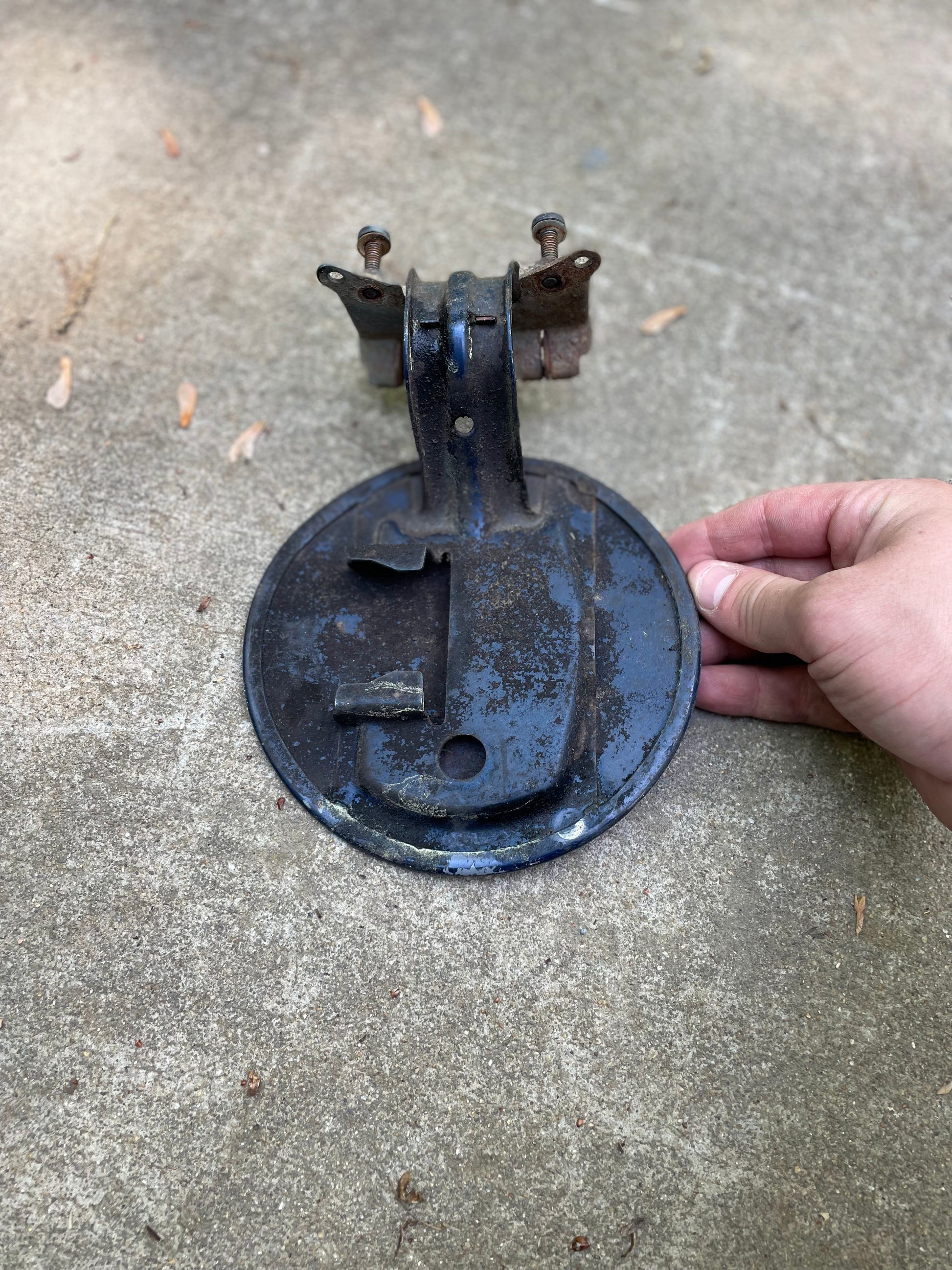 A hand holds a weathered blue OEM Fuel Filler Neck Door Gas Tank Cover by General Motors for 2001-2004 Chevy S10 & GMC Sonoma, showing rust and wear, with bracket and bolts attached, resting on concrete.