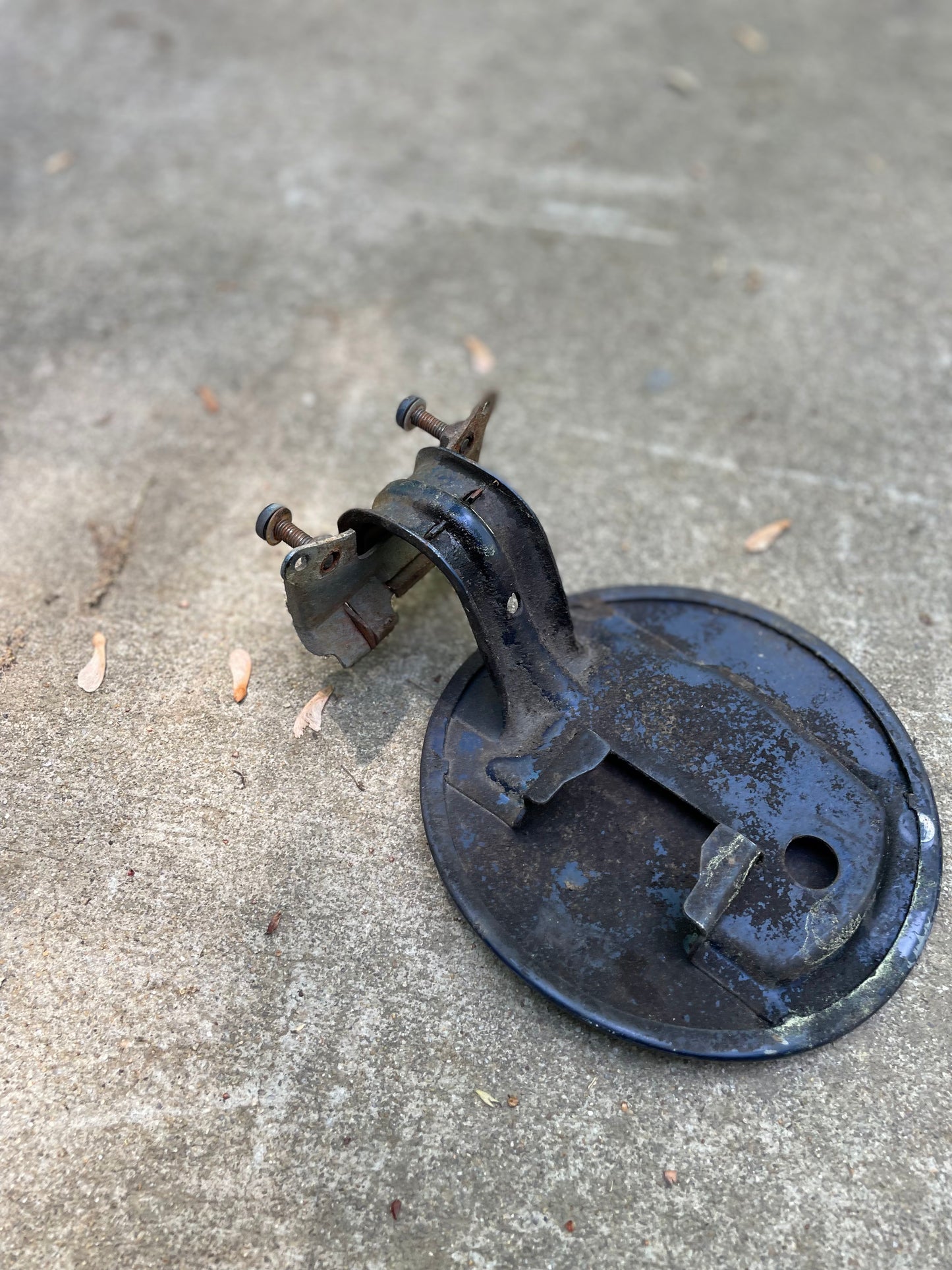 A blue General Motors OEM fuel filler neck door gas tank cover for 2001-2004 Chevy S10 & GMC Sonoma lies on a concrete surface, its attached bracket visible and surrounded by scattered leaves.