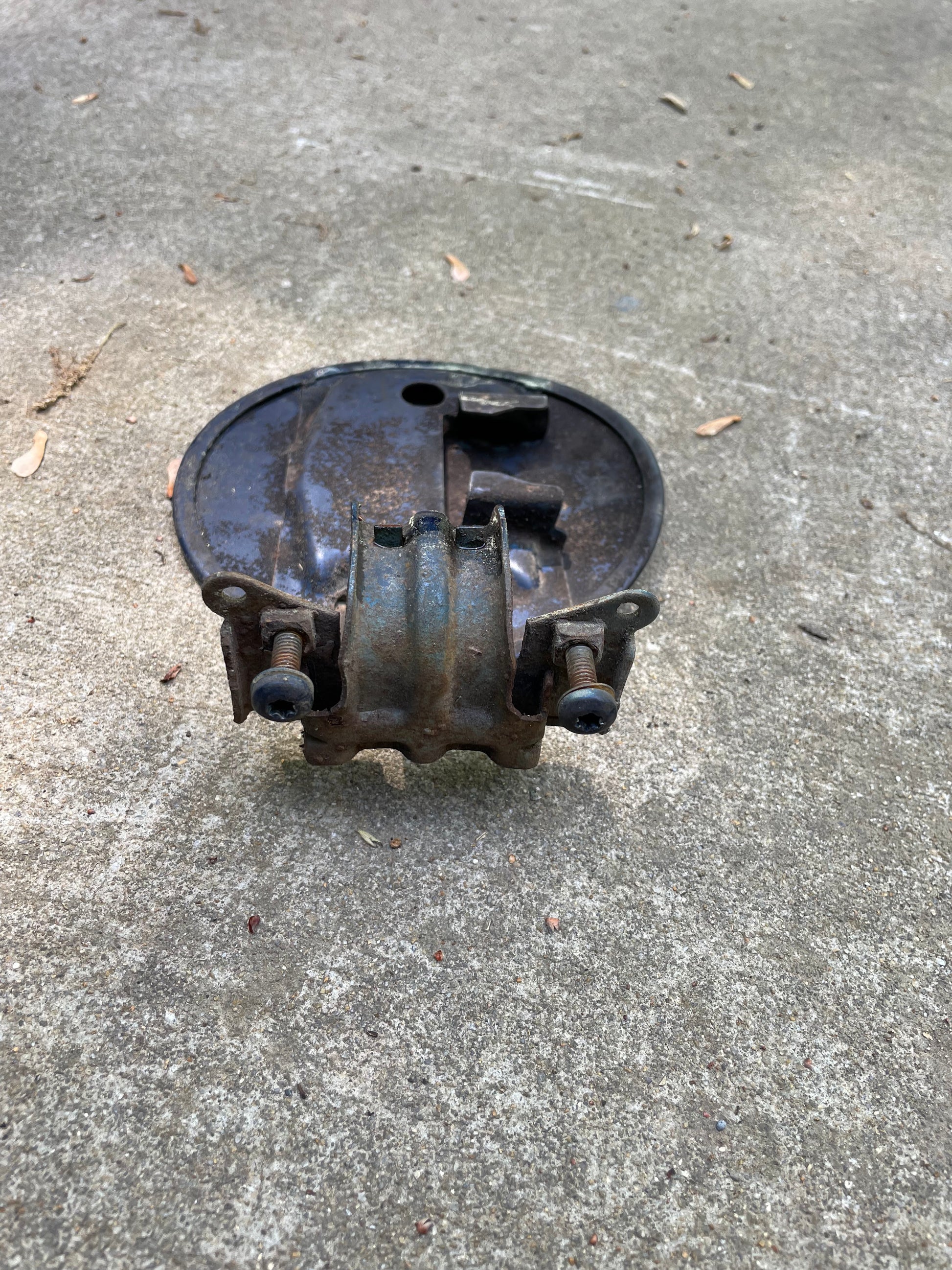 A weathered General Motors OEM Fuel Filler Neck Door Gas Tank Cover in Blue for 2001-2004 Chevy S10 & GMC Sonoma, showing rust and corrosion, rests outdoors on rough concrete.