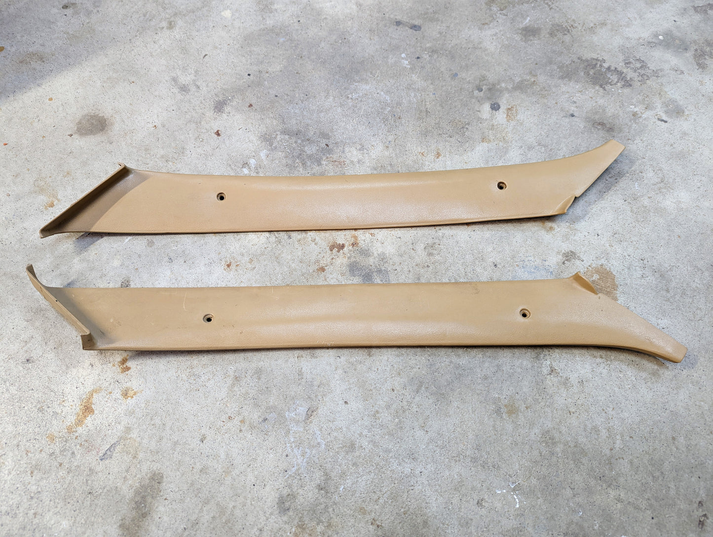Two tan OEM General Motors A Pillar Trim Panels (64I) for 1982-1993 Chevrolet S10, Blazer, and more, shown parallel with screw holes on a concrete floor—ideal originals for restoration or upgrade.