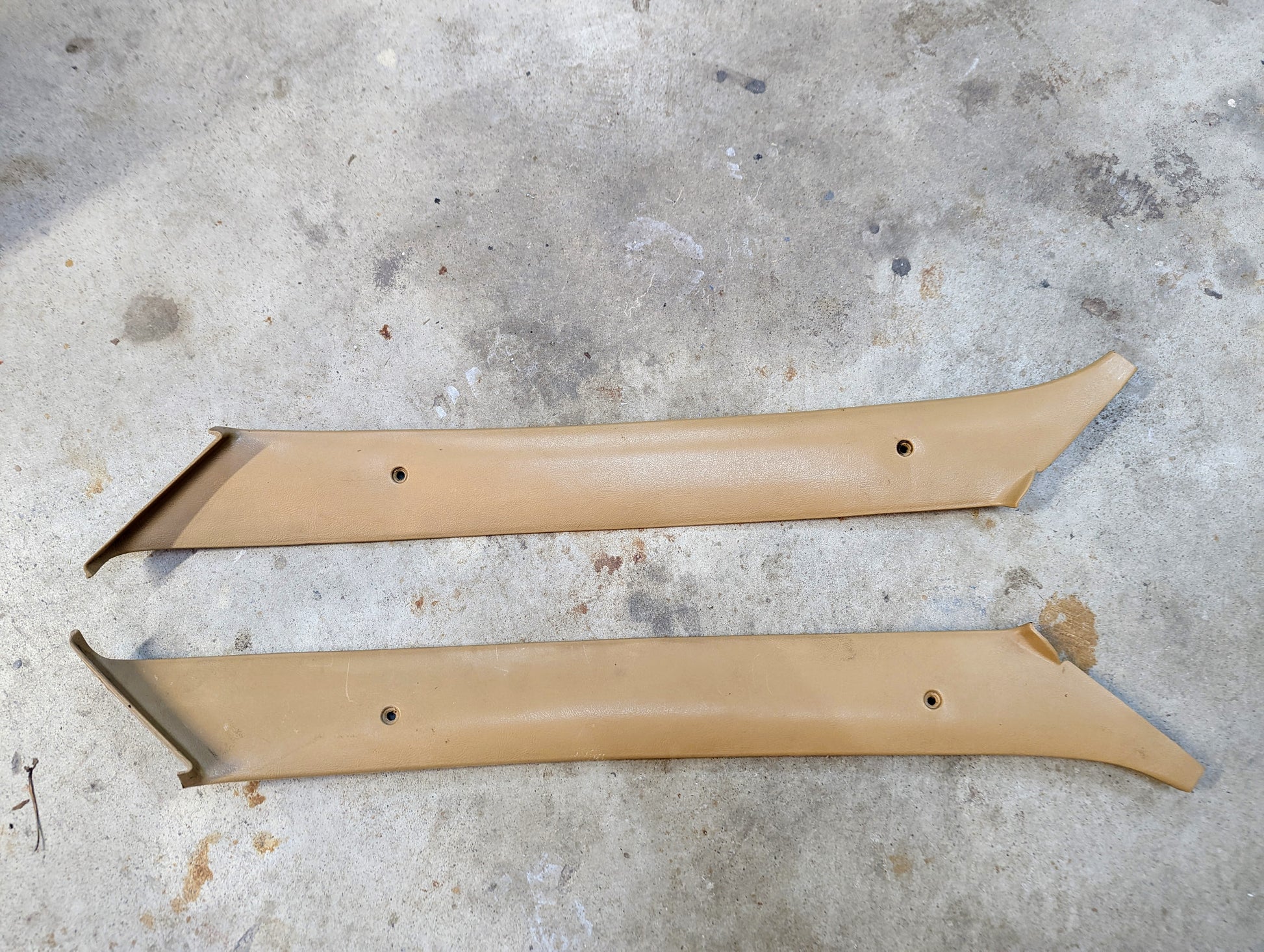 A pair of tan OEM A Pillar Trim Panels (64I) by General Motors for 1982–1993 Chevrolet S10, Blazer, and more, showing slight wear, are shown on a concrete surface—ideal for restoring your vehicle’s interior.