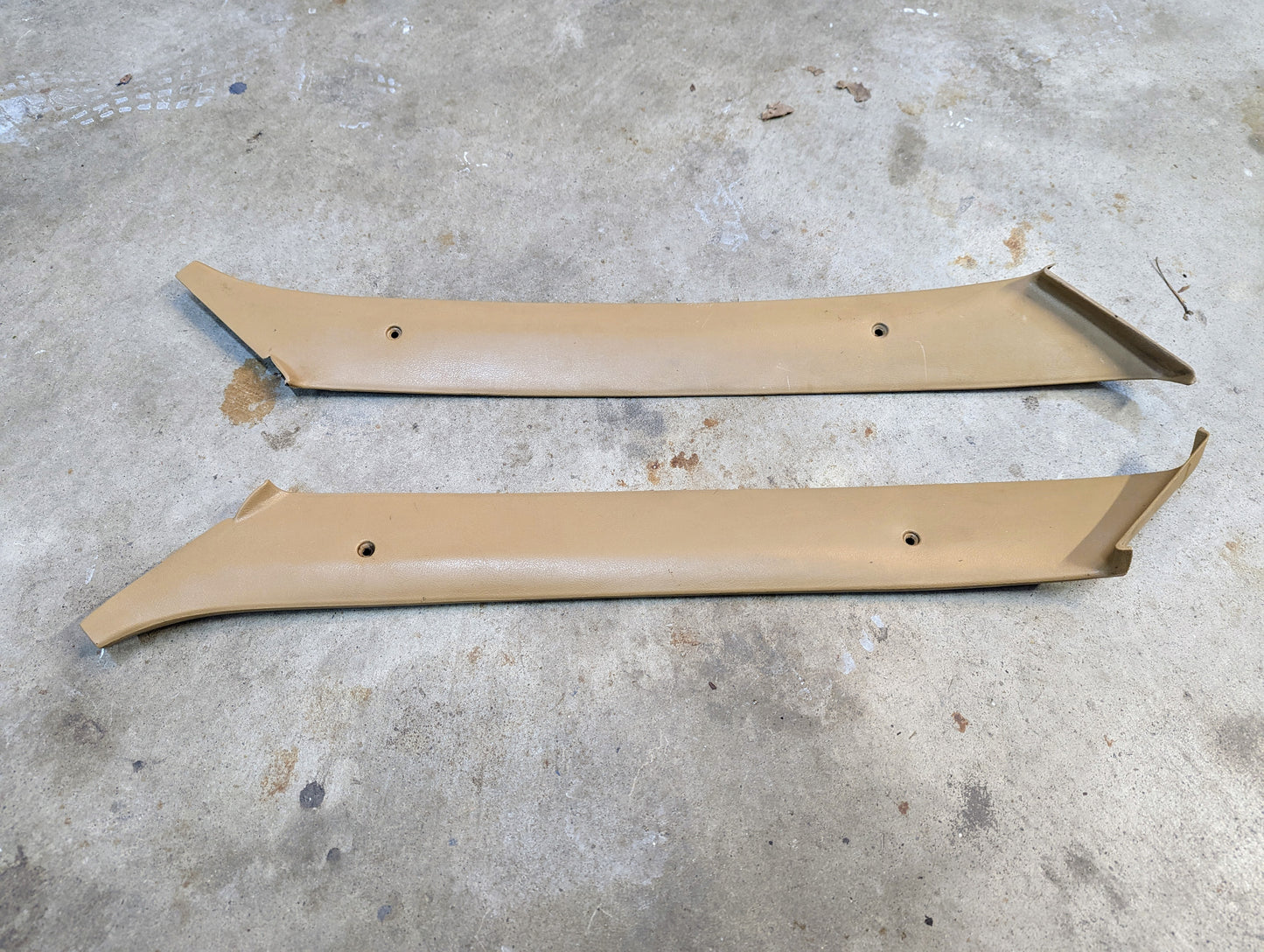 A pair of tan OEM A Pillar Trim Panels (64I) by General Motors for 1982–1993 Chevrolet S10, Blazer, and more, shown with screw holes and angled ends, lying parallel on a concrete floor.