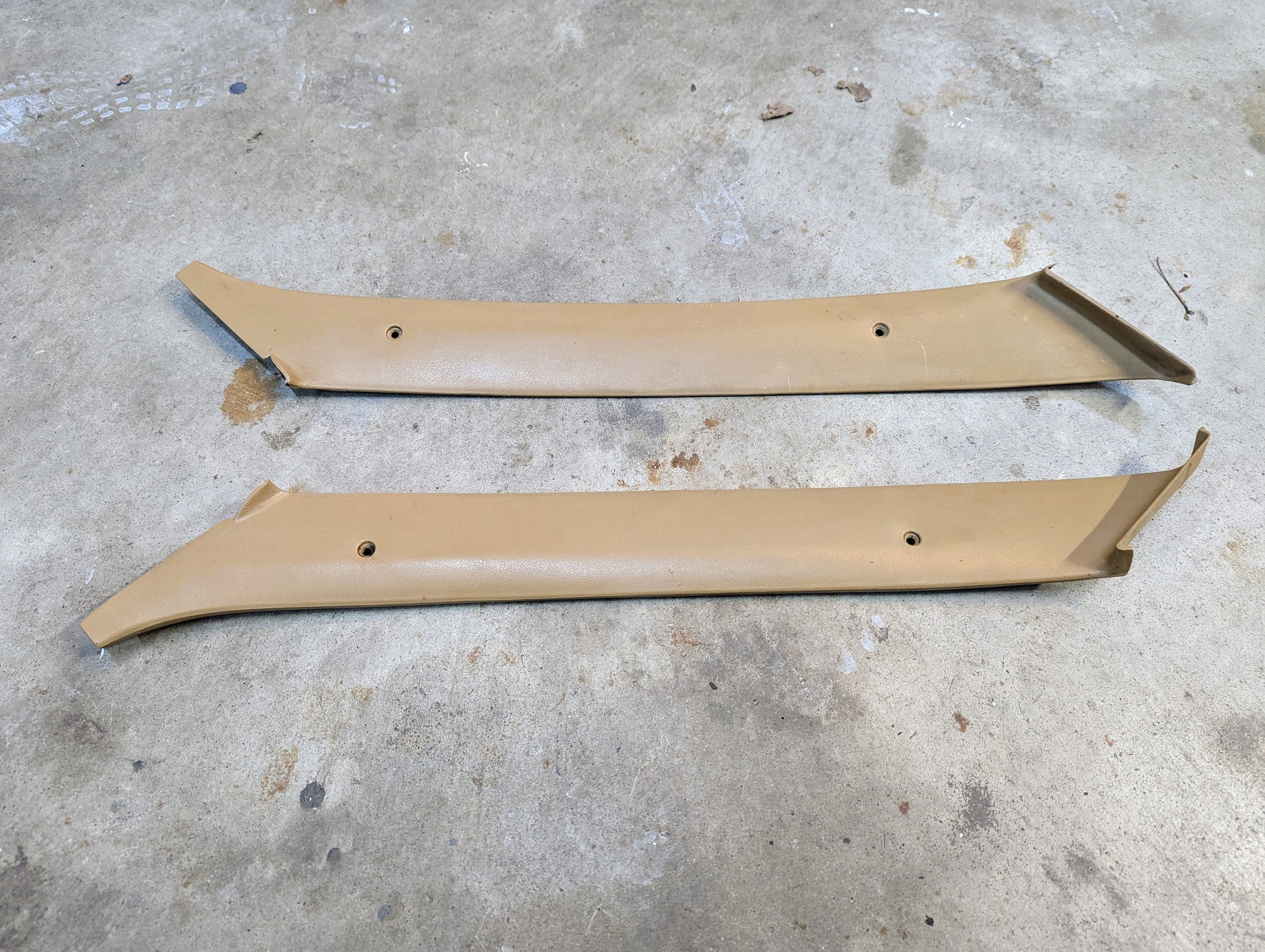 A pair of tan OEM A Pillar Trim Panels (64I) by General Motors for 1982–1993 Chevrolet S10, Blazer, and more, shown with screw holes and angled ends, lying parallel on a concrete floor.