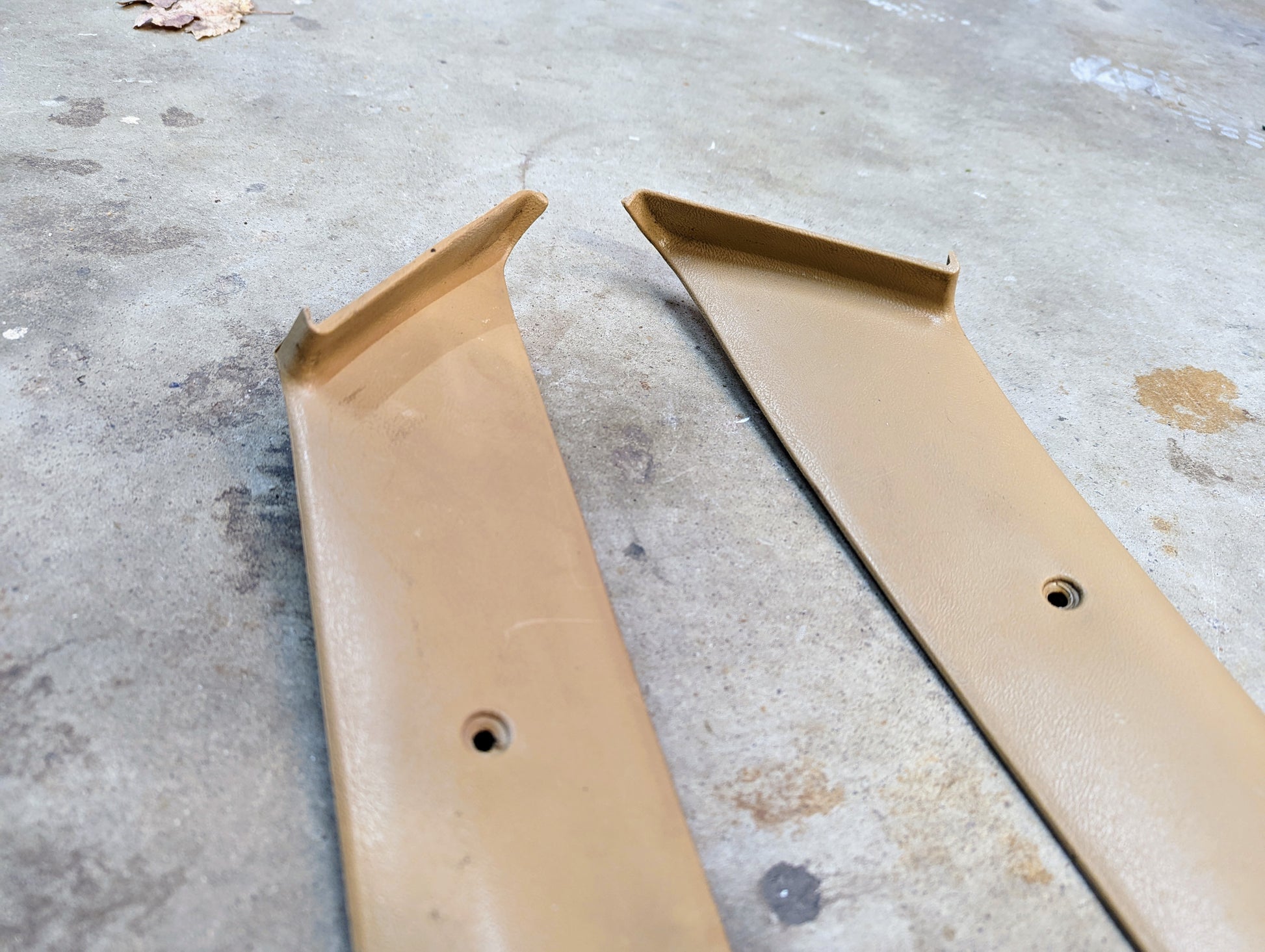 A pair of tan OEM A Pillar Trim Panels (64I) for 1982-1993 Chevrolet S10, Blazer, and more by General Motors are shown with angled ends and screw holes on a concrete surface, positioned parallel and displaying slight wear.