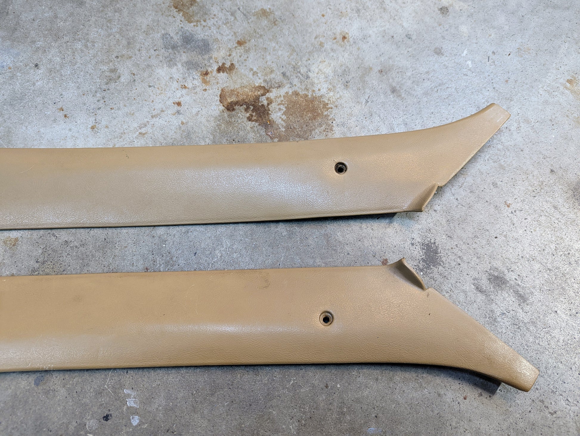 A pair of OEM General Motors A Pillar Trim Panels in Tan 64I for 1982-1993 Chevrolet S10, Blazer, and others, with screw holes and some end damage, are shown on a gray stained concrete floor.