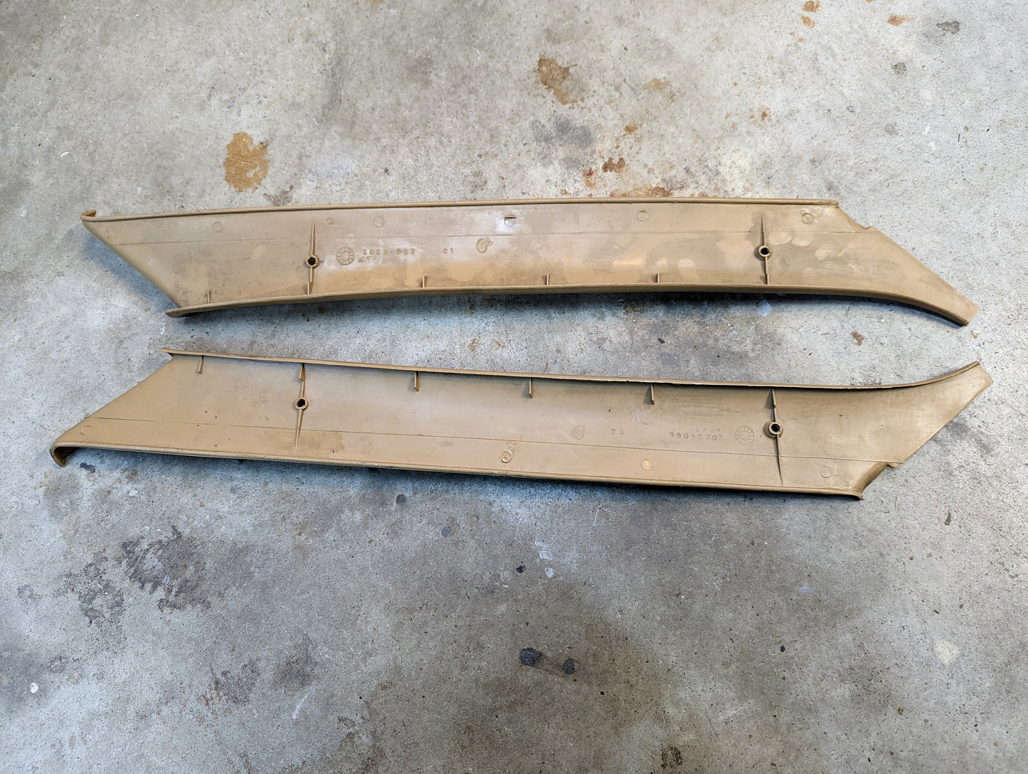 A pair of tan OEM General Motors A Pillar Trim Panels (64I) for 1982–1993 Chevrolet S10, Blazer, and more, lying parallel on a concrete floor with visible mounting points and minor surface scratches.