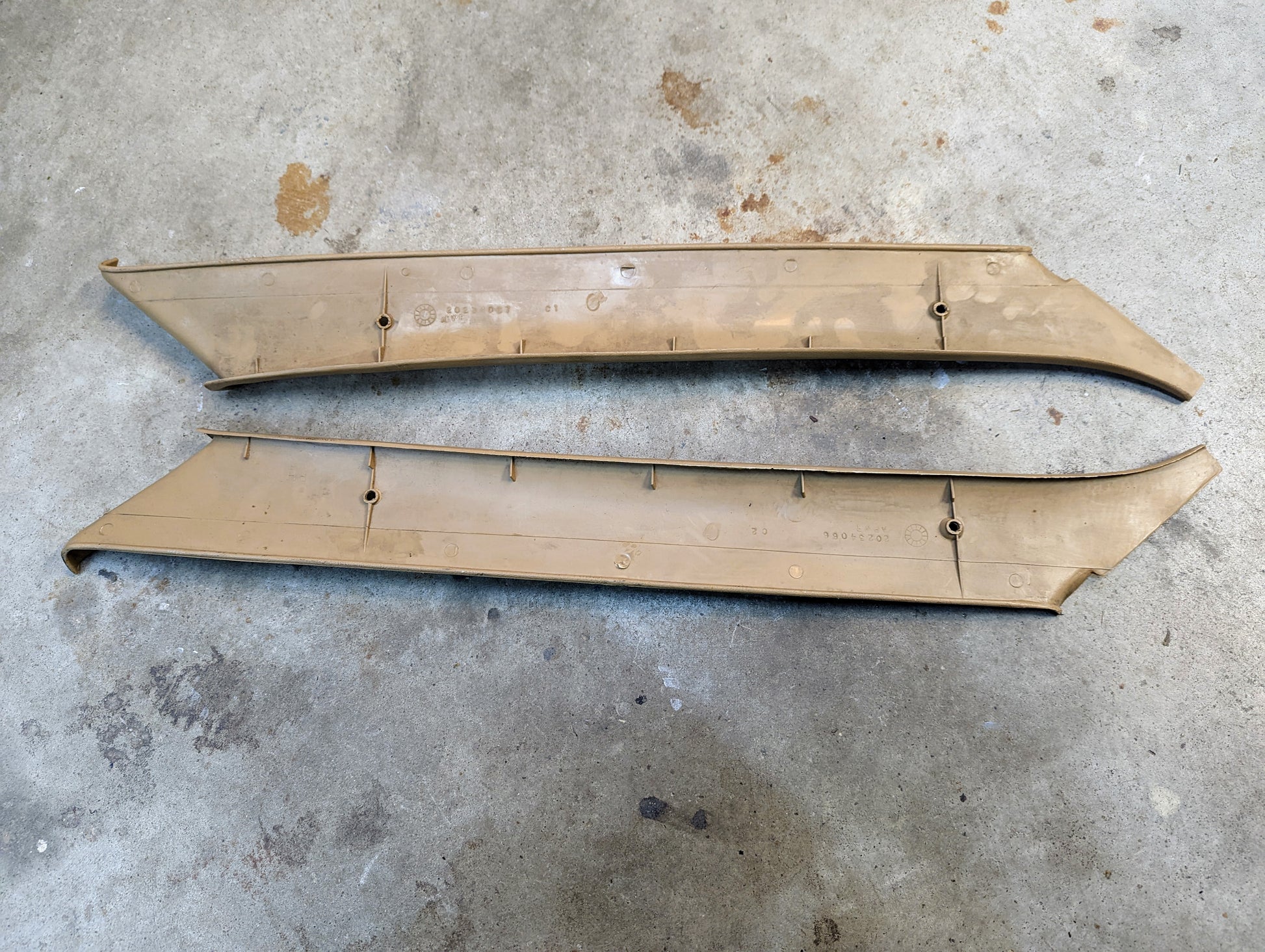 A pair of tan OEM General Motors A Pillar Trim Panels (64I) for 1982–1993 Chevrolet S10, Blazer, and more, lying parallel on a concrete floor with visible mounting points and minor surface scratches.
