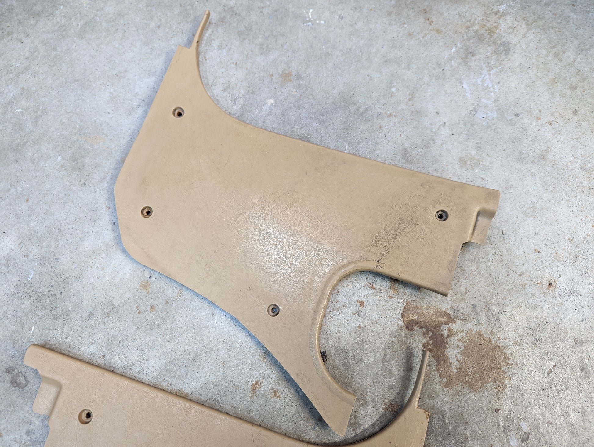 A General Motors OEM Pair Kick Panel Knee Trim in Tan 64I for 1982-1993 Chevrolet S10, Blazer, and more lies on gray concrete, showing screw holes, wear, and a curved cutout. Made for Beige 64I trim.