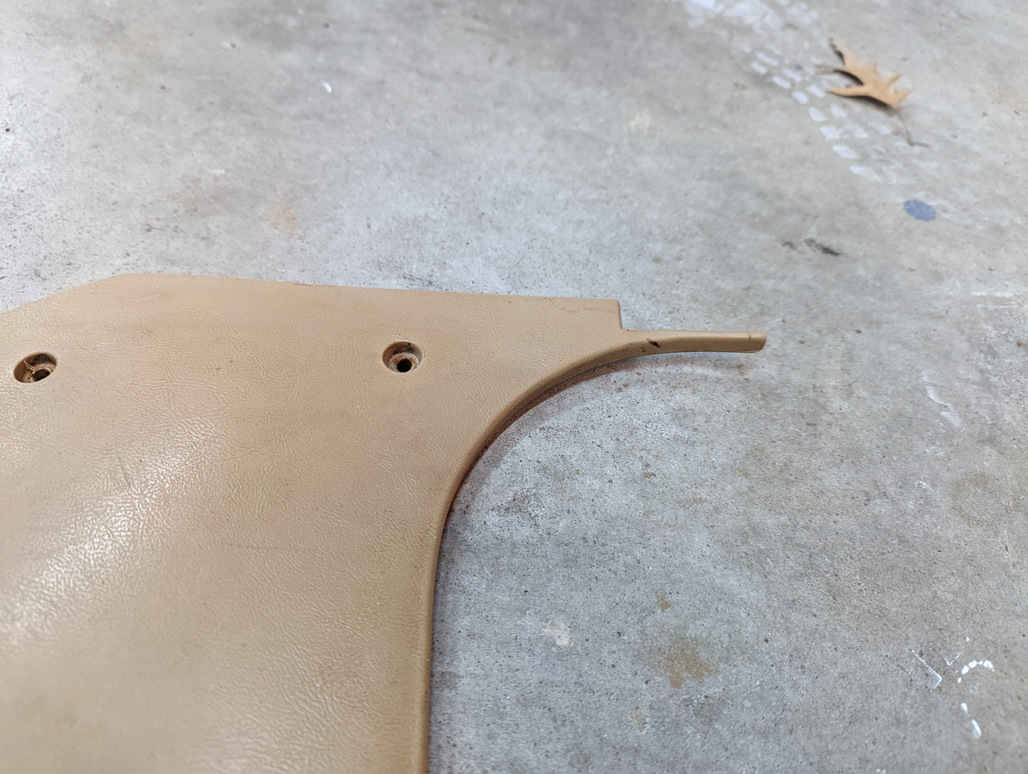 A tan OEM Pair Kick Panel Knee Trim 64I by General Motors for 1982-1993 Chevrolet S10/Blazer lies on a gray floor, showing two screw holes and a jagged edge. A small brown leaf sits in the top right corner.