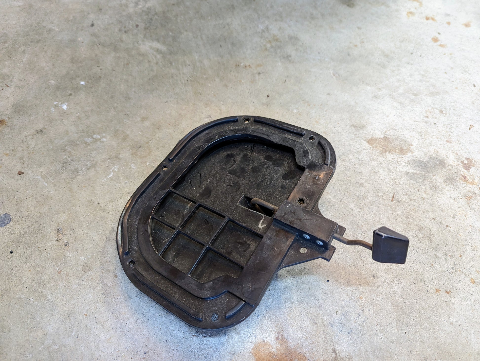 A pair of OEM Fresh Air Floor Vent Doors by General Motors for 1982-1993 Chevrolet S10, Blazer, and more, is lying on a concrete floor; the black rectangular metal parts appear worn with some rust and dirt visible.