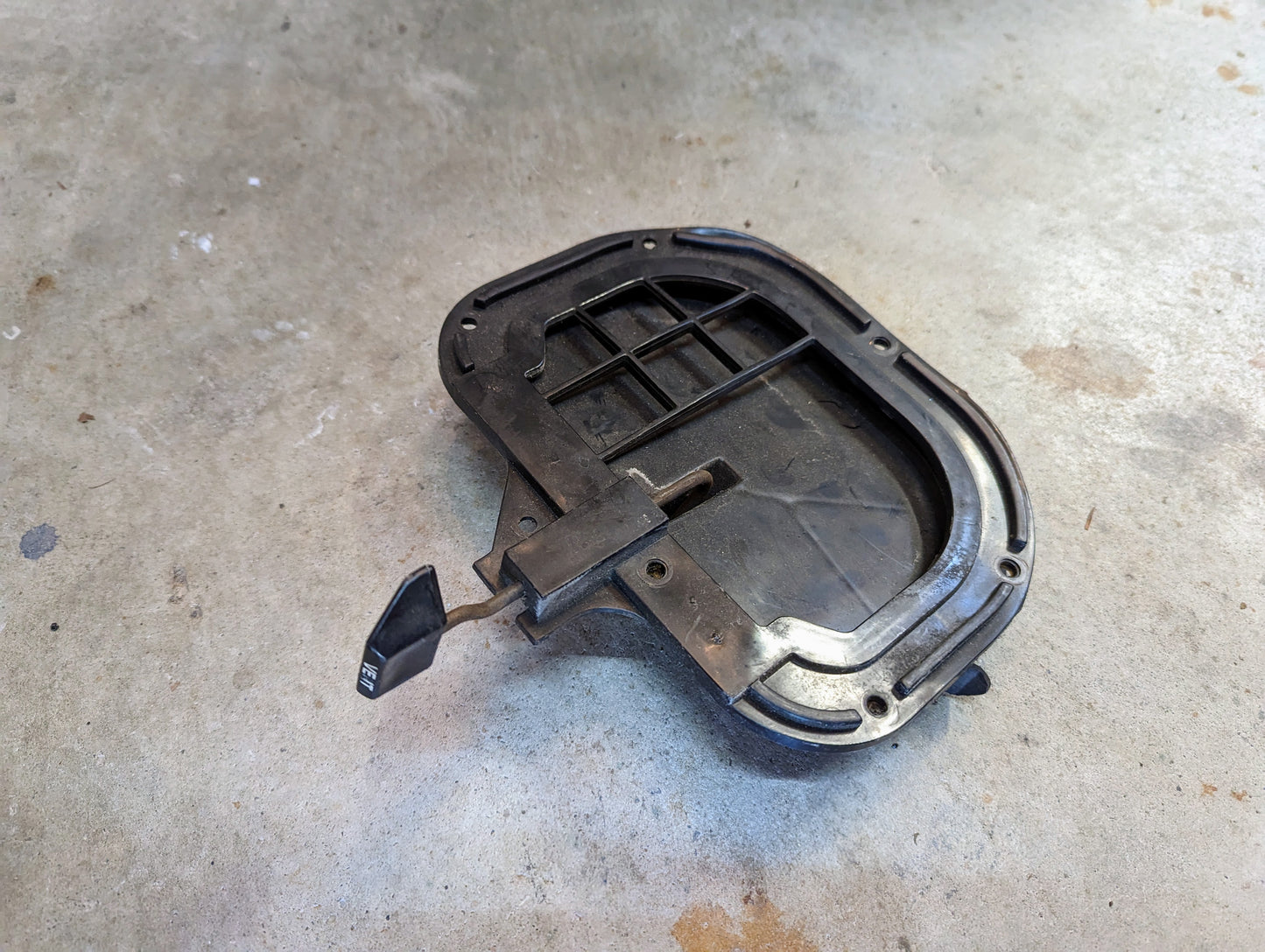 A black plastic OEM Fresh Air Floor Vent Door Pair by General Motors, with lever and labels, lies on concrete showing dirt and use—fits 1982-1993 Chevrolet S10, Blazer, and more; features curved edges and internal supports.