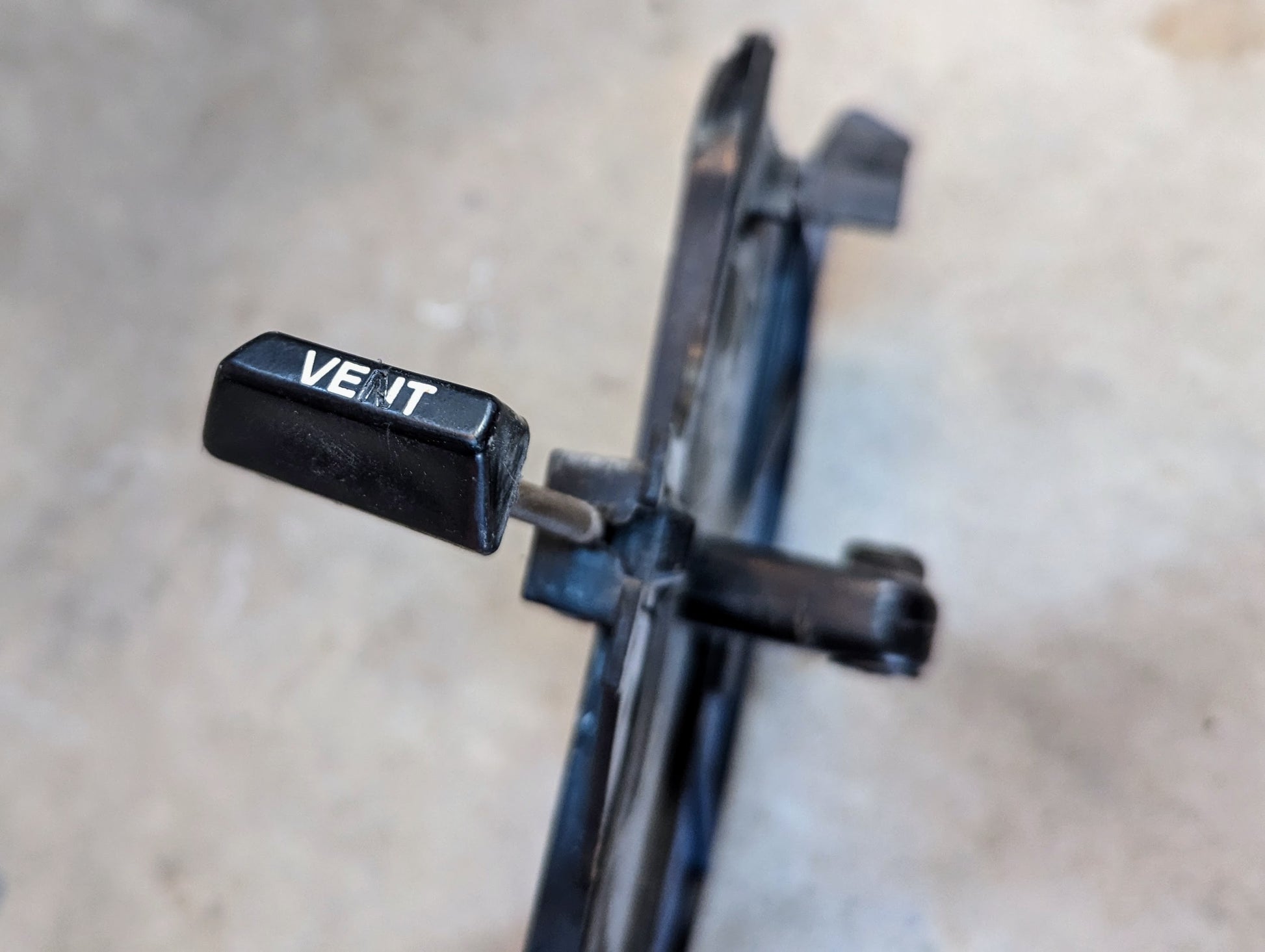 A close-up of a black lever labeled VENT on a General Motors OEM Fresh Air Floor Vent Door Pair for 1982-1993 Chevrolet S10, Blazer, and more, shown attached to the metal mechanism with a blurred gray background.