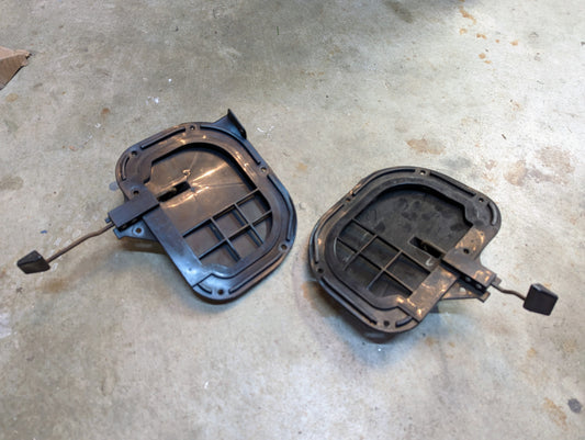 A pair of OEM General Motors Fresh Air Floor Vent Doors for 1982-1993 Chevrolet S10, Blazer, and others, rest on concrete. The black plastic vents with attached metal rods show dirt and wear from previous use.