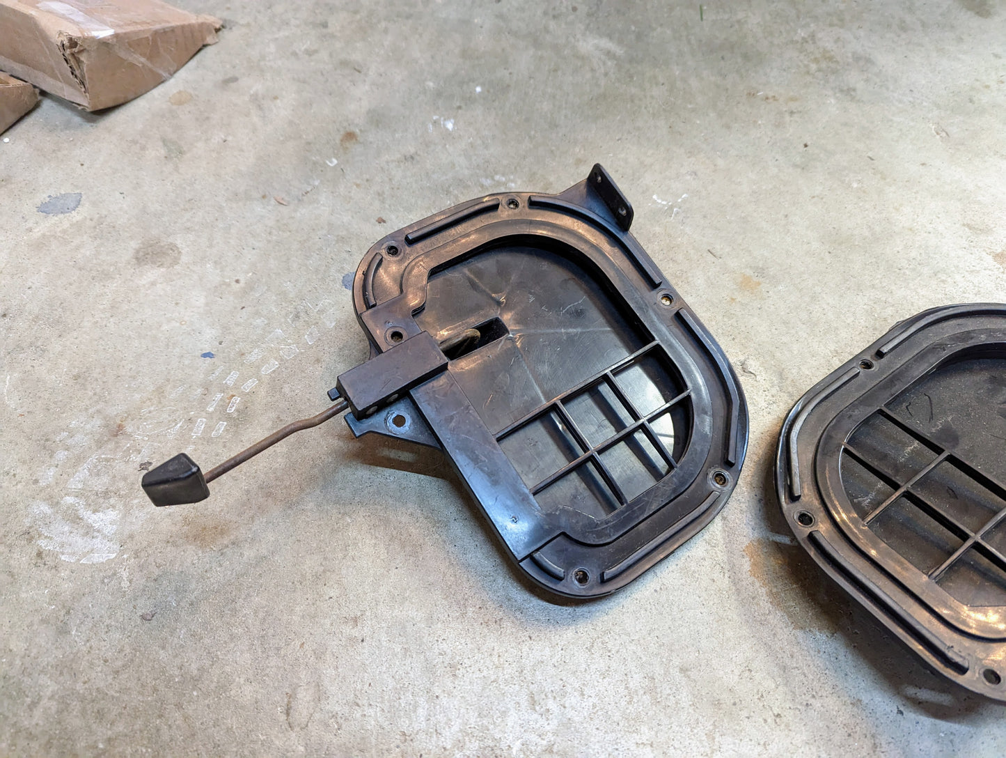 A General Motors OEM Fresh Air Floor Vent Door Pair for 1982-1993 Chevrolet S10, Blazer, and more lies on a concrete floor; one vent door is fully visible, another partially shown. A cardboard box sits in the upper left corner.