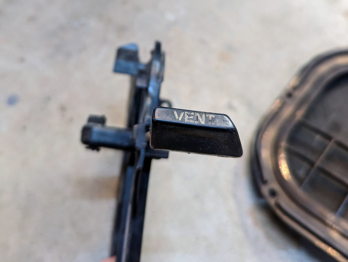 Close-up of a black plastic lever labeled VENT, attached to a General Motors OEM Fresh Air Floor Vent Door (pair) for 1982-1993 Chevrolet S10, Blazer, and more, with a blurred background and part of another black object on the right.