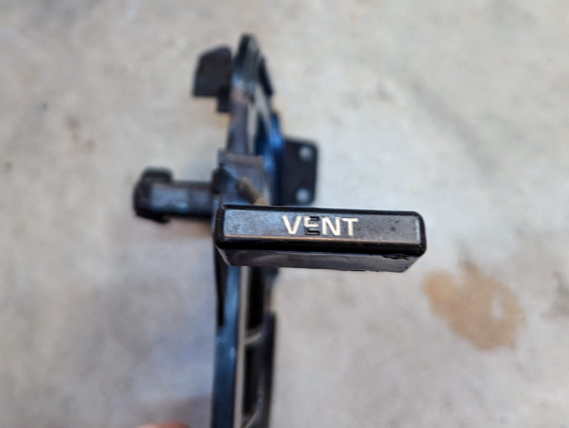 A close-up of a black driver-side OEM Fresh Air Floor Vent Door by General Motors for 1982–1993 Chevrolet S10, Blazer, and more, with VENT printed on the end, set against a blurred light background.