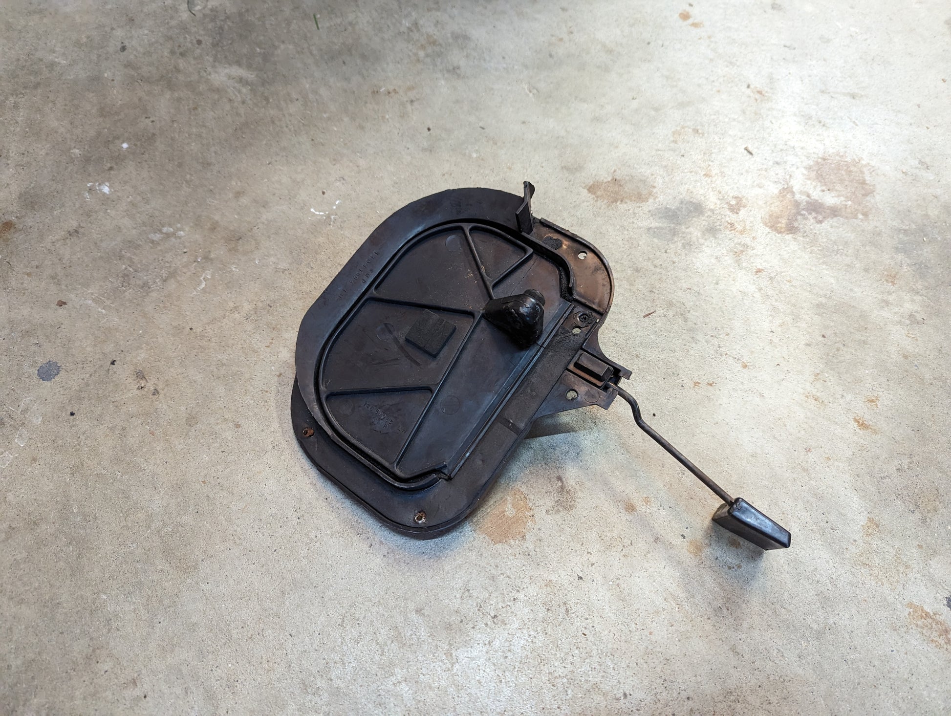 A worn, slightly bent OEM Fresh Air Floor Vent Door (Driver-Side LH) by General Motors for 1982-1993 Chevrolet S10 and Blazer features a lever and latch; its seen here resting on a concrete floor.
