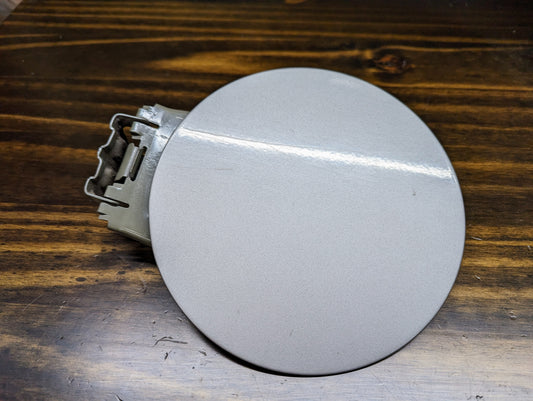 A round, gray OEM Fuel Filler Neck Door Gas Tank Cover in Silver Birch for 1999-2006 Chevy Silverado & GMC Sierra by General Motors rests on a dark wooden surface, featuring an attached metal hinge.
