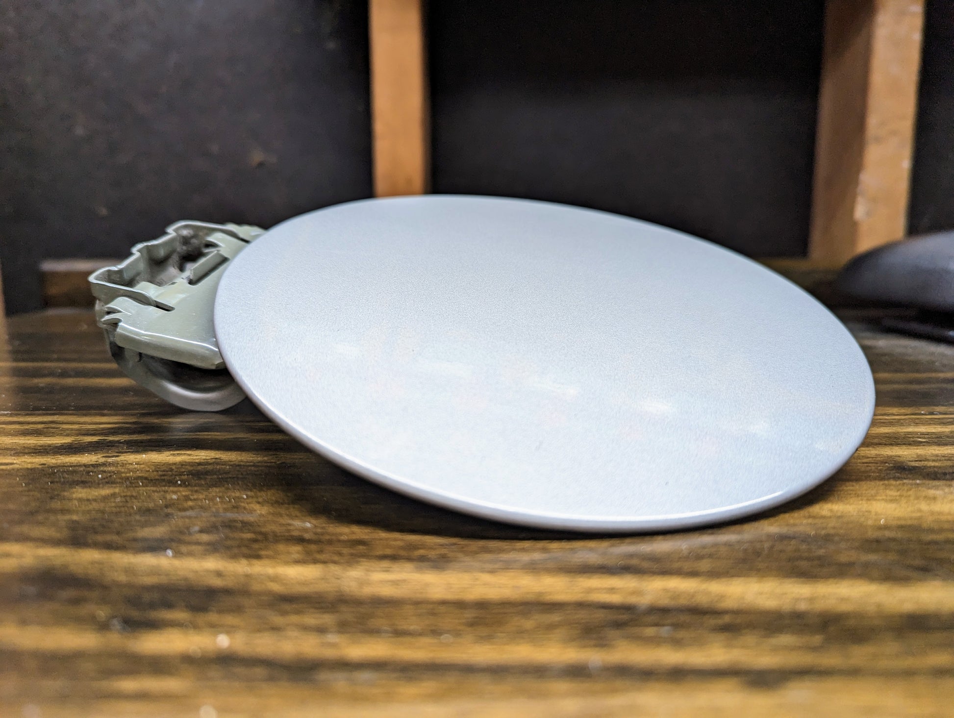 A General Motors OEM Fuel Filler Neck Door Gas Tank Cover in Silver Birch for 1999-2006 Chevy Silverado & GMC Sierra, with hinge attached, rests on a wooden surface against a dark blurred background.