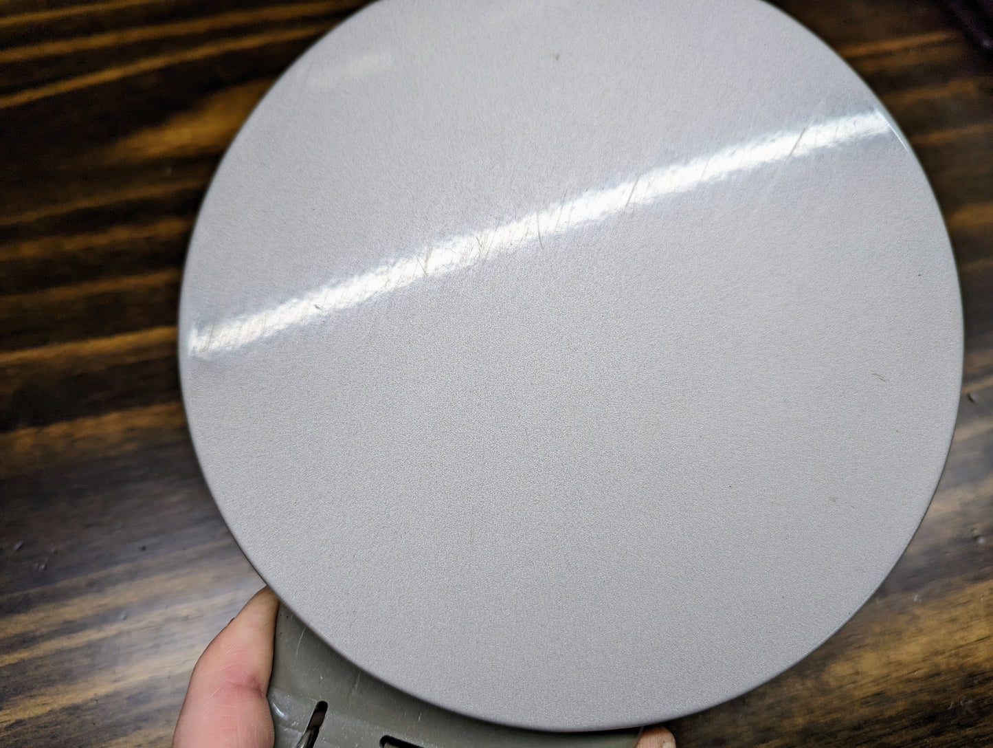 A close-up of a round, smooth General Motors OEM Fuel Filler Neck Door Gas Tank Cover in Silver Birch for 1999-2006 Chevy Silverado & GMC Sierra, held by hand over a dark wood background, with faint scratches and bright light reflection.