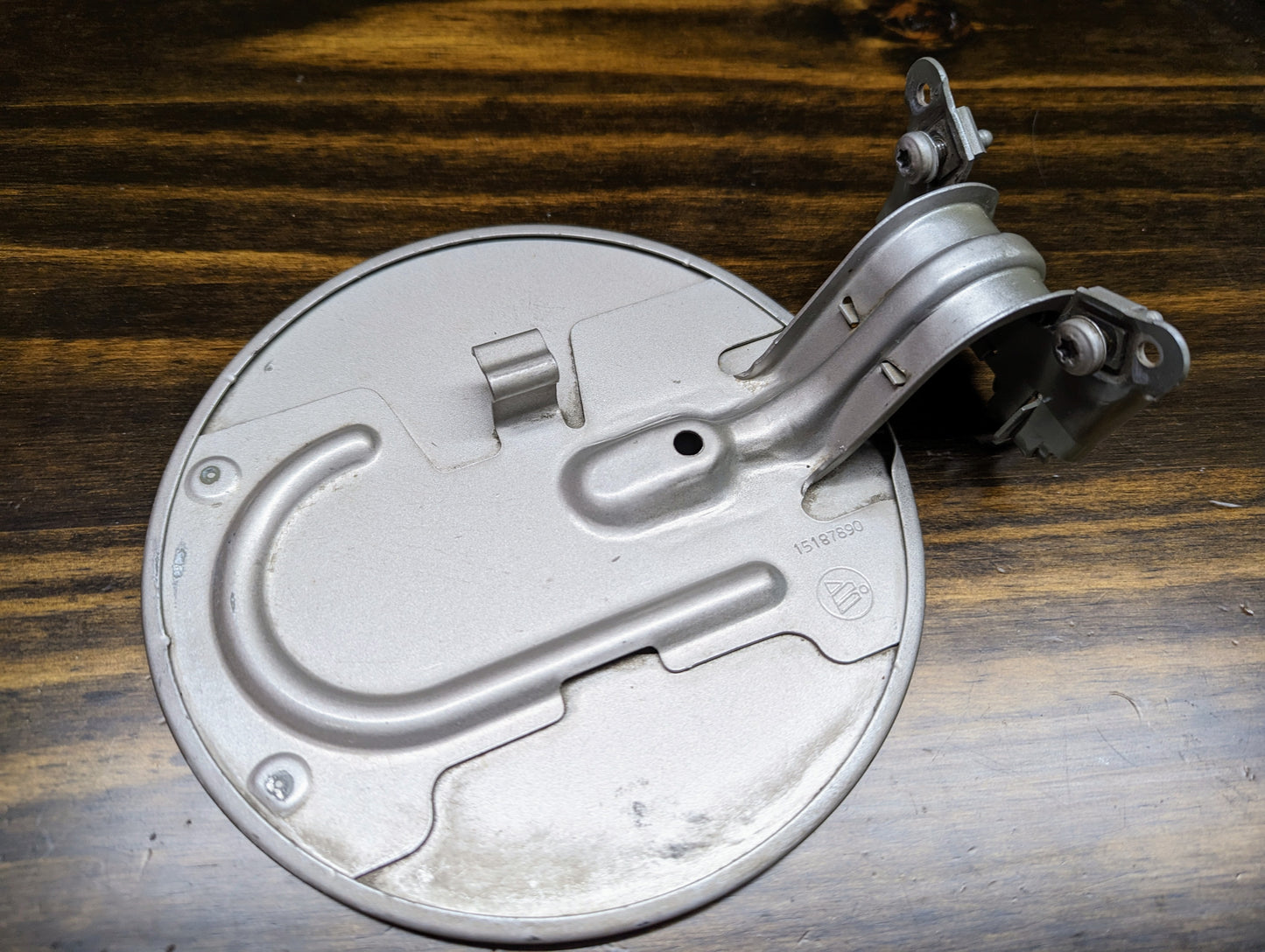An OEM Fuel Filler Neck Door Gas Tank Cover in Silver Birch for 1999-2006 Chevy Silverado & GMC Sierra by General Motors, featuring a hinge, visible screws, and curved indentation, displayed on a dark wooden surface.