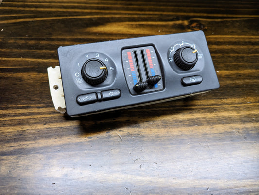 A General Motors OEM Dual Climate HVAC Manual Control Head w/Rear Defrost for 2003-2006 GM Full Size Trucks is shown on a wooden surface, featuring rotary dials and sliders for fan speed, temperature, and airflow direction.