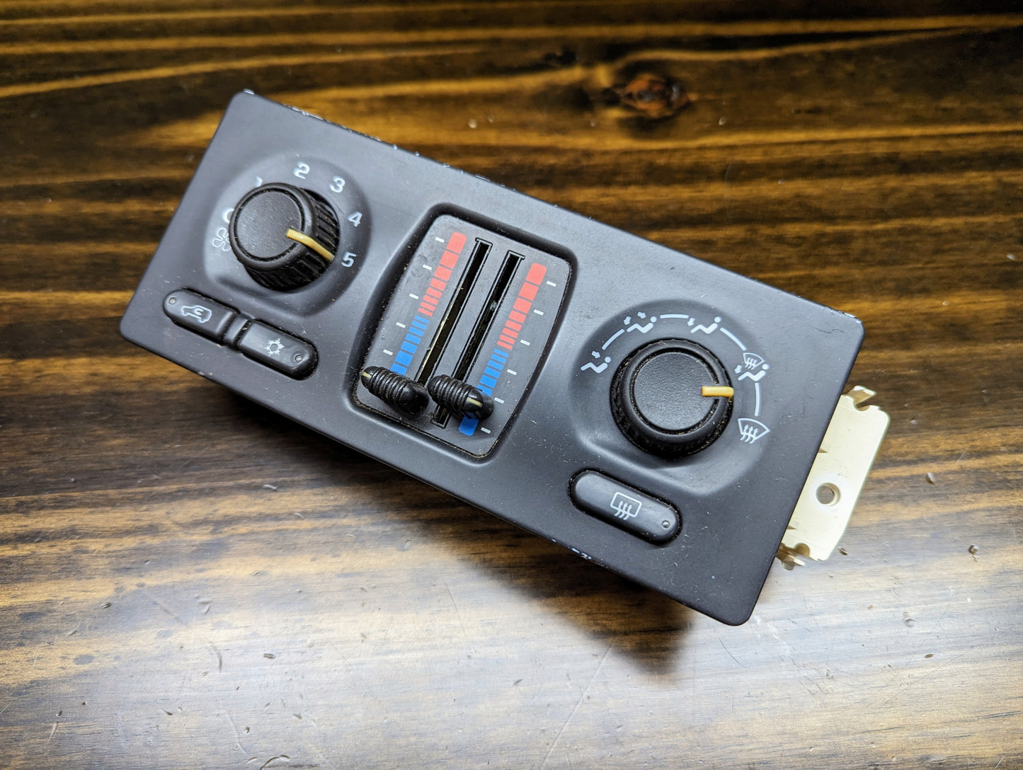 A close-up of the OEM Dual Climate HVAC Manual Control Head with Rear Defrost for 2003-2006 GM Full Size Trucks by General Motors, showing temperature sliders, fan speed dial, air direction settings, and rear defrost button on a wooden surface.