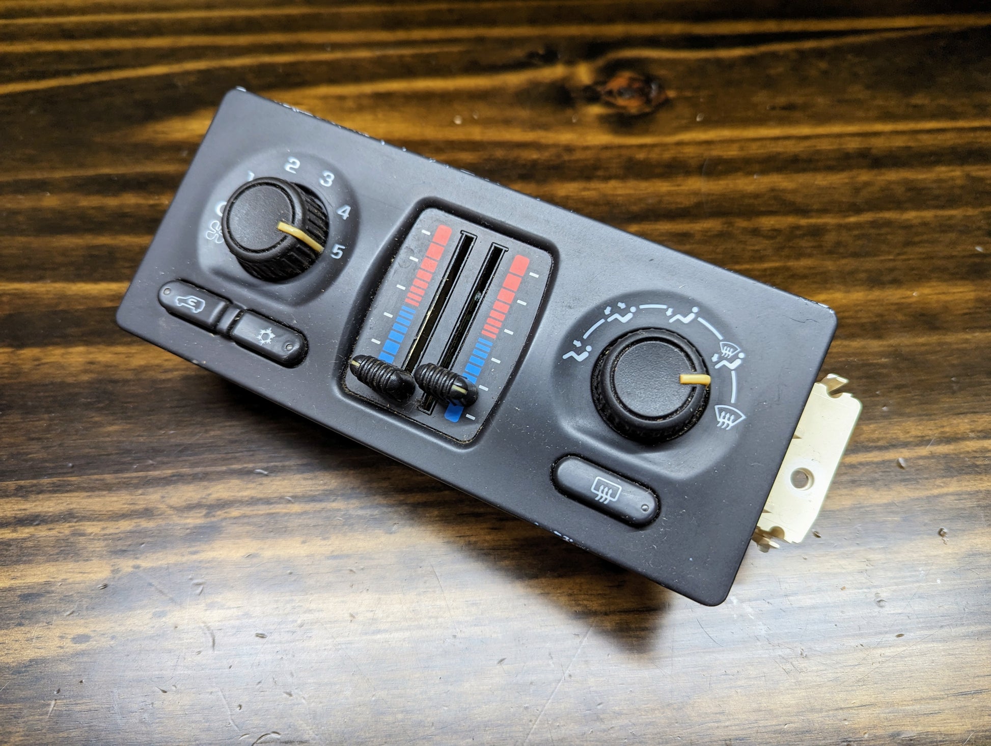 A close-up of the OEM Dual Climate HVAC Manual Control Head with Rear Defrost for 2003-2006 GM Full Size Trucks by General Motors, showing temperature sliders, fan speed dial, air direction settings, and rear defrost button on a wooden surface.