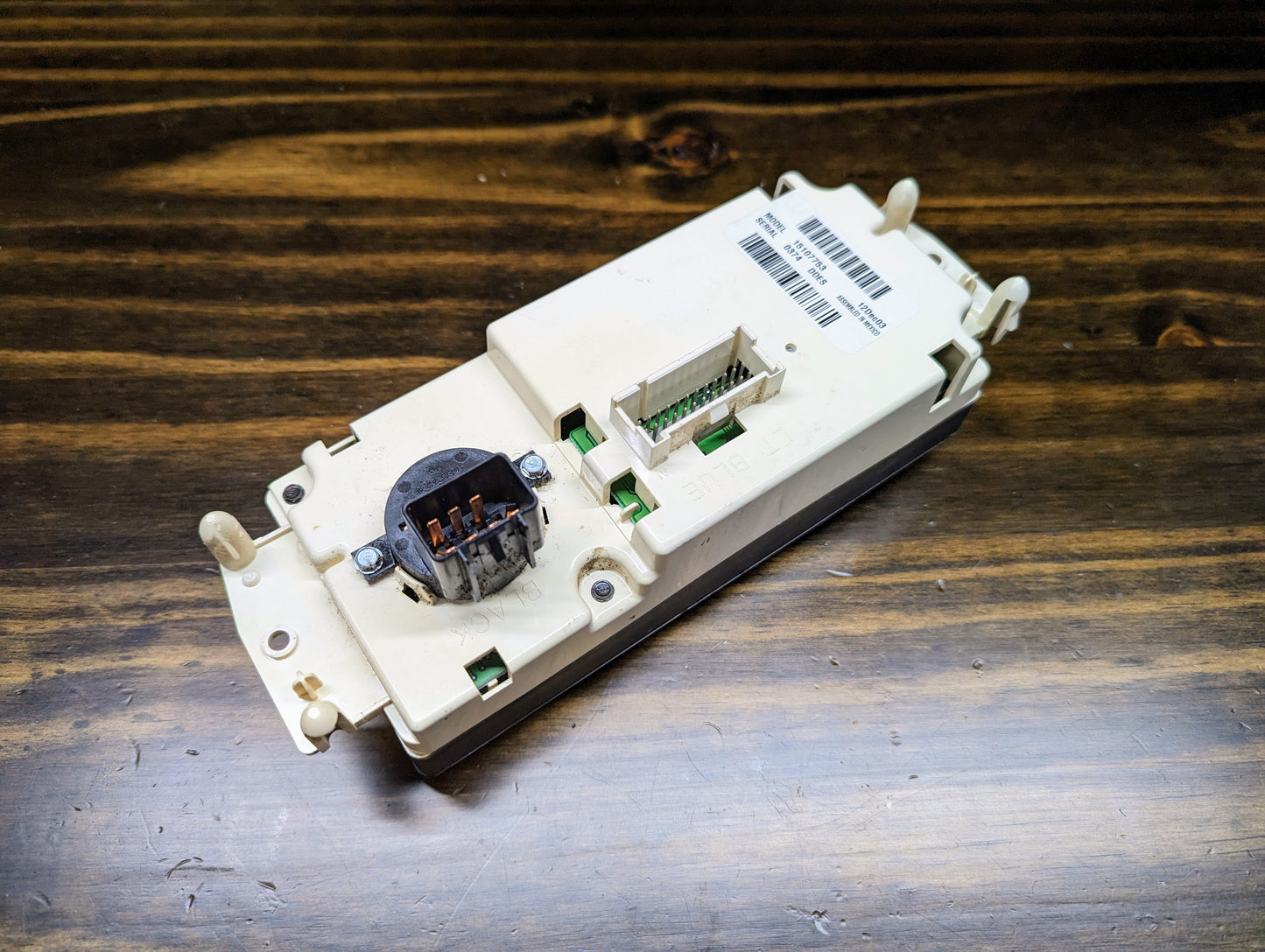 A close-up of the General Motors OEM Dual Climate HVAC Manual Control Head with Rear Defrost for 2003-2006 GM Full Size Trucks, showing its connectors, labels, and barcodes, resting on a wooden surface.