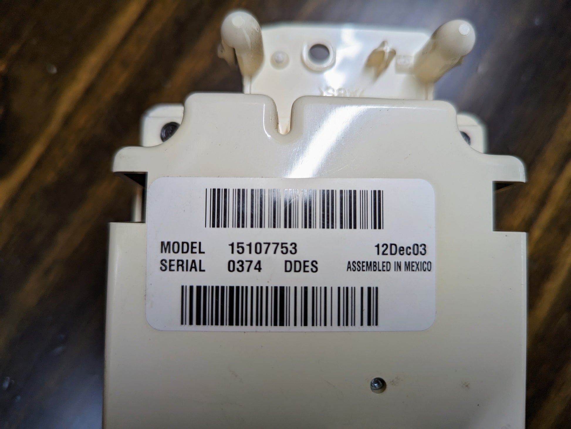 Close-up of a General Motors OEM Dual Climate HVAC Manual Control Head w/Rear Defrost for 2003–2006 GM Full Size Trucks, showing barcode label MODEL 15107753, SERIAL 0374 DDES, 12Dec03, ASSEMBLED IN MEXICO on a dark wooden surface.