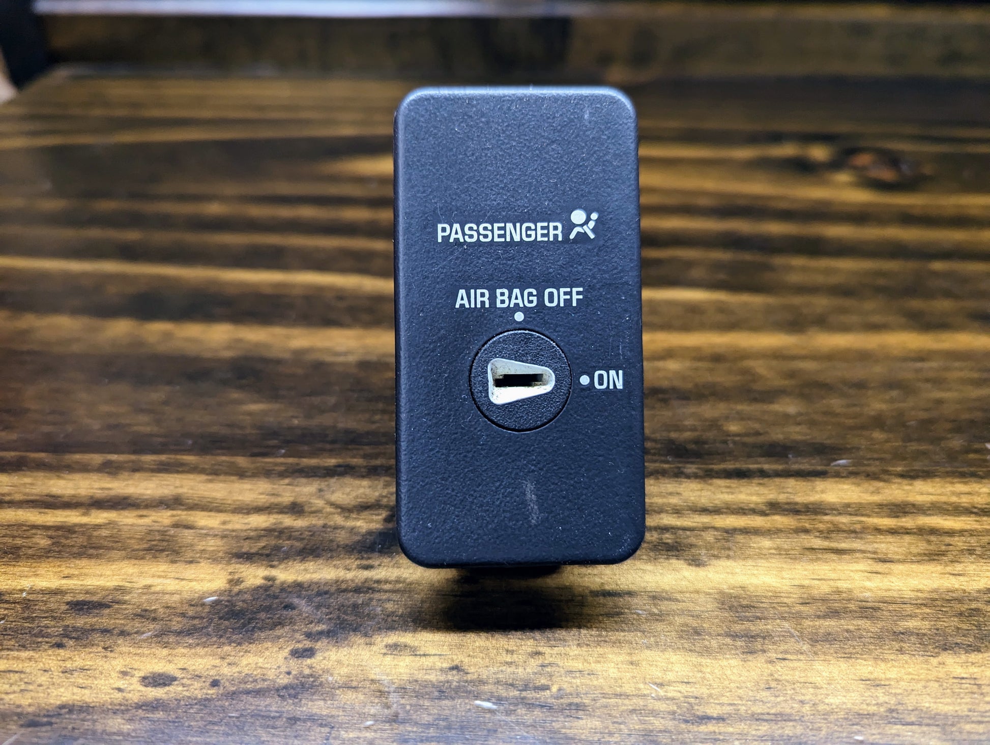 A close-up of a General Motors OEM Passenger Inflatable Restraint Air Bag Key Switch for 1998-2003 Chevy S10 & GMC Sonoma, showing the PASSENGER AIR BAG OFF label, key slot, and ON indicator on a wooden surface.