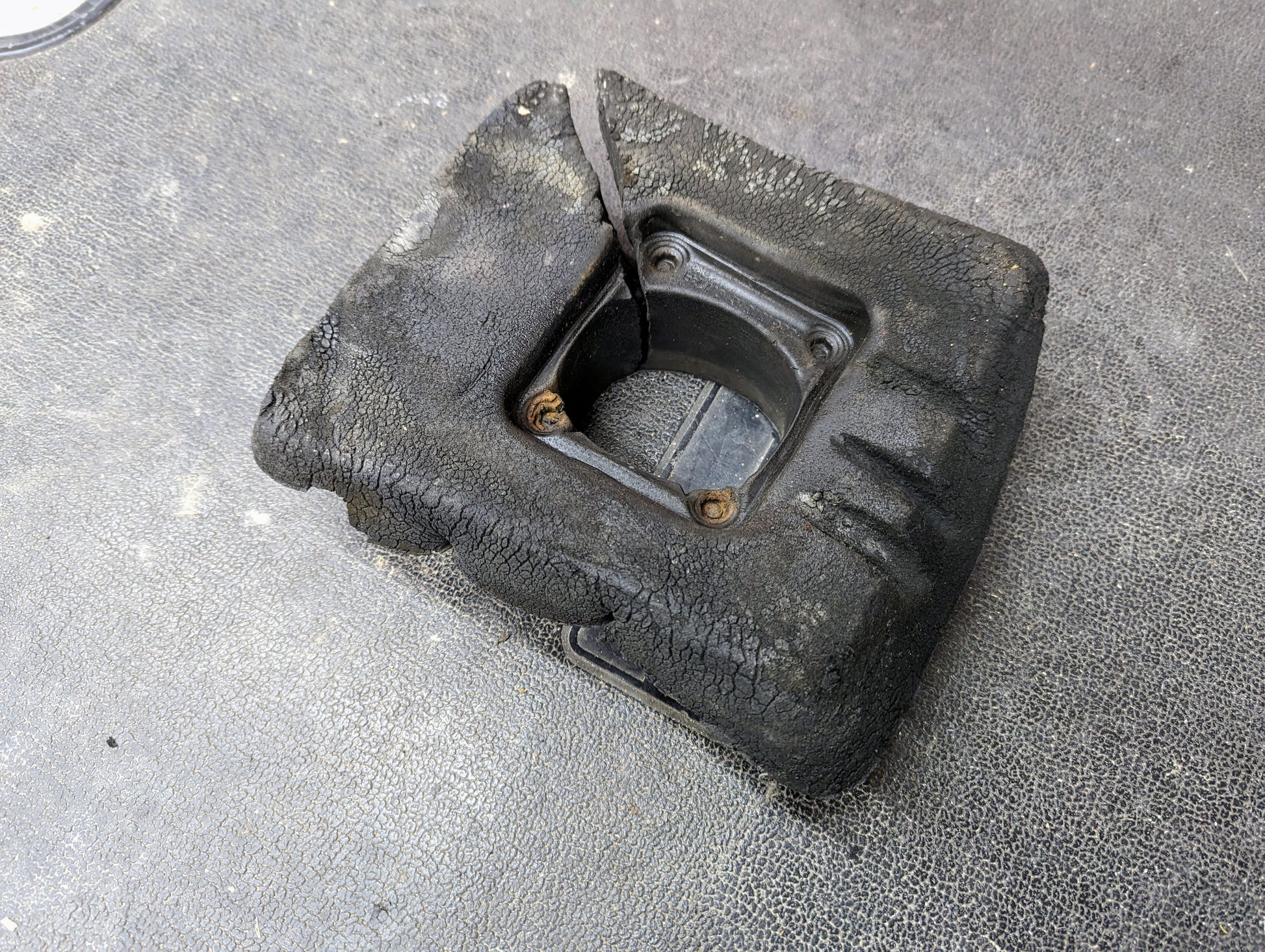 A worn General Motors Manual Floor Shifter Insulator Foam for 1998-2005 Chevy S10 Blazer GMC Sonoma Jimmy lies on a textured dark surface. Four screws hold the center, with visible cracks and a partly detached rubber strip.
