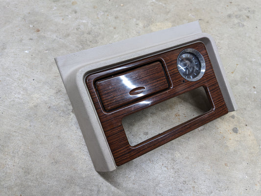 A General Motors OEM Center Console Front Bin Woodgrain Trim with Clock in Light Beige for 2003-2006 Cadillac Escalade is shown placed on a concrete surface.