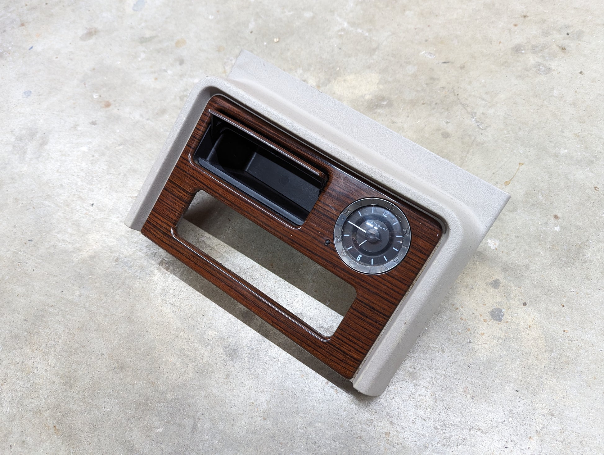 A General Motors OEM Center Console Front Bin Woodgrain Trim with Clock in Light Beige for 2003-2006 Cadillac Escalade rests on a concrete floor.