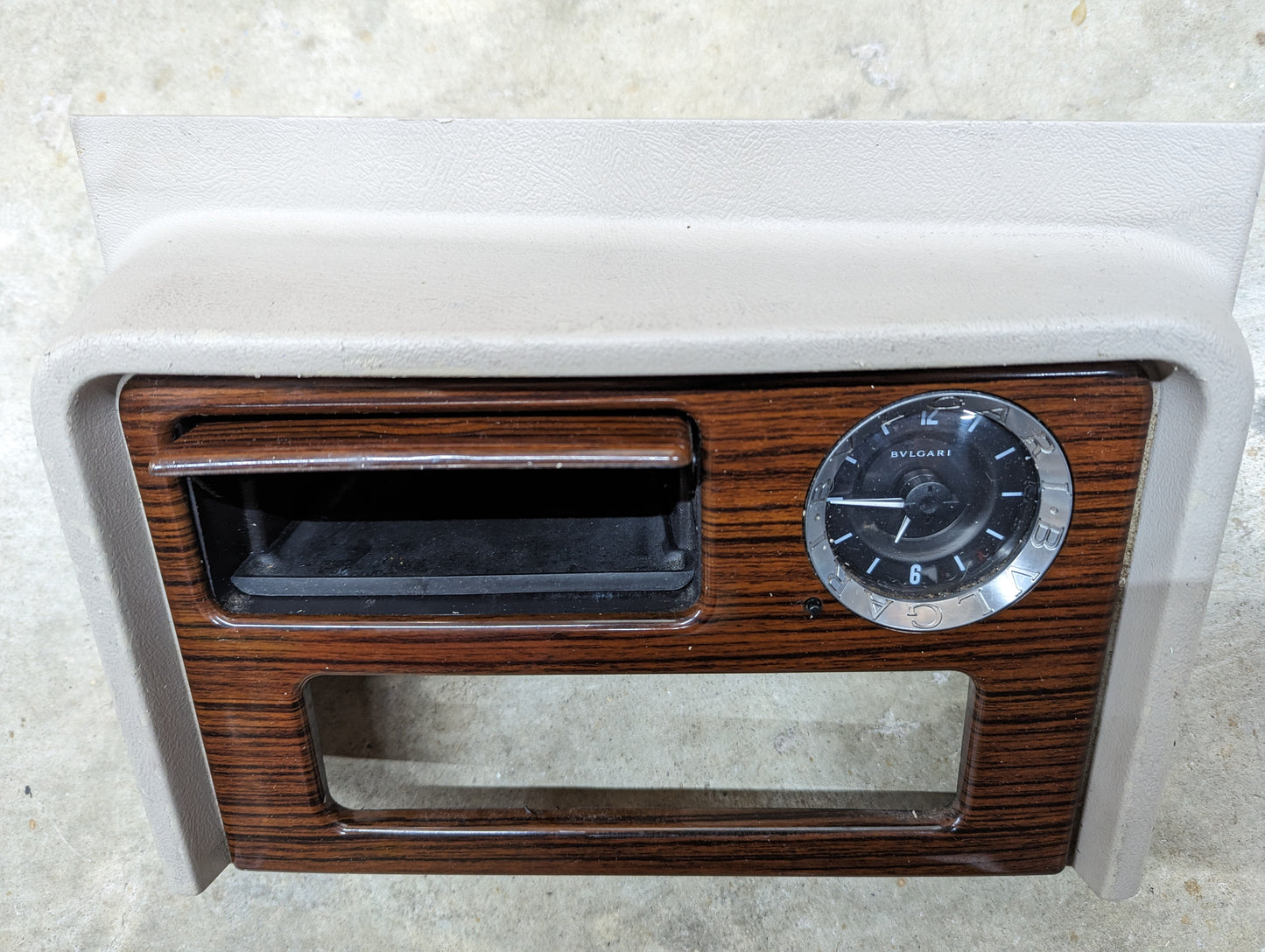 The General Motors OEM Center Console Front Bin Woodgrain Trim with Clock in Light Beige fits 2003-2006 Cadillac Escalade, featuring a vintage look with an analog clock, center slot, and lower opening—ideal for restoring your interior.