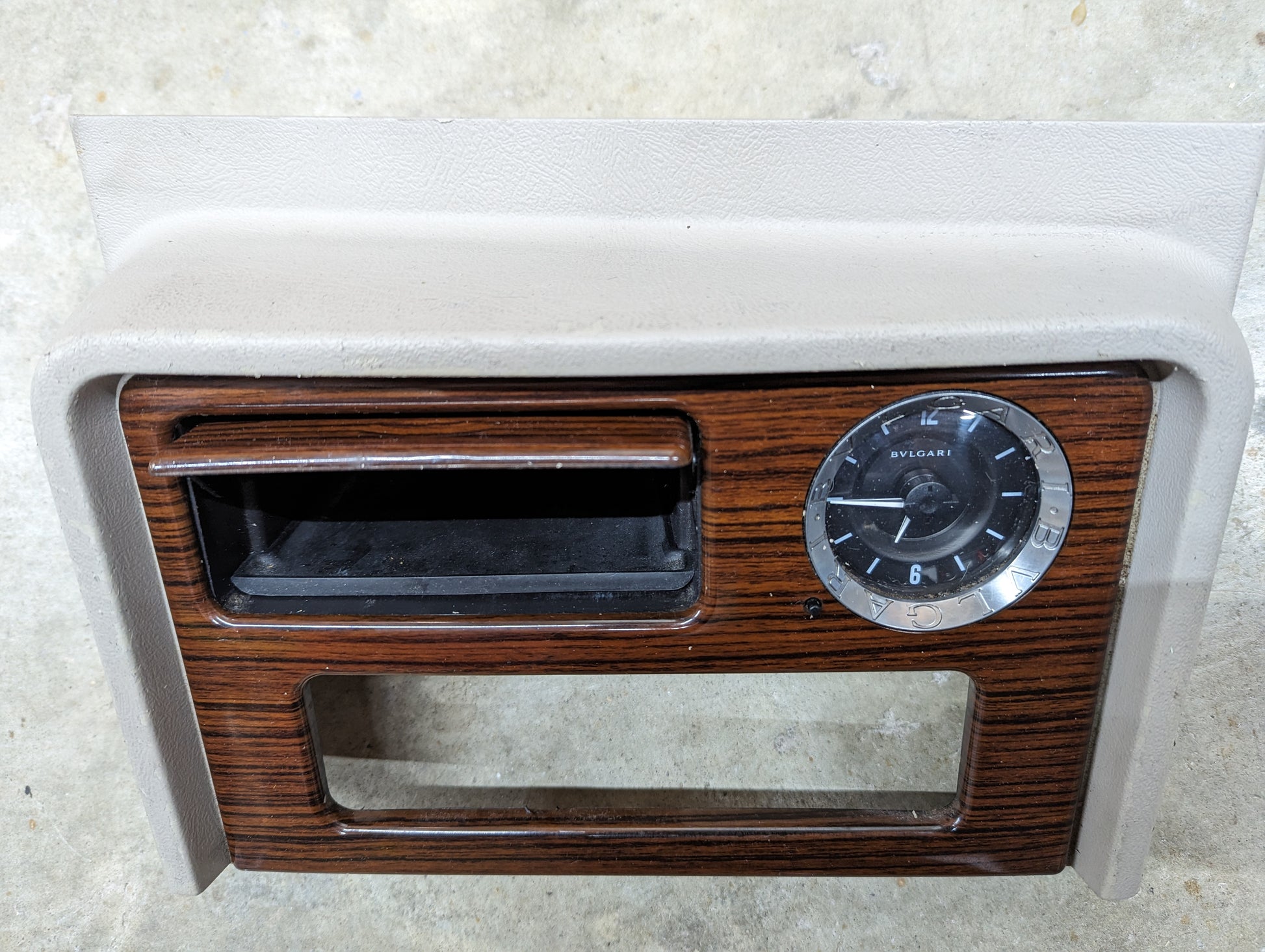 The General Motors OEM Center Console Front Bin Woodgrain Trim with Clock in Light Beige fits 2003-2006 Cadillac Escalade, featuring a vintage look with an analog clock, center slot, and lower opening—ideal for restoring your interior.