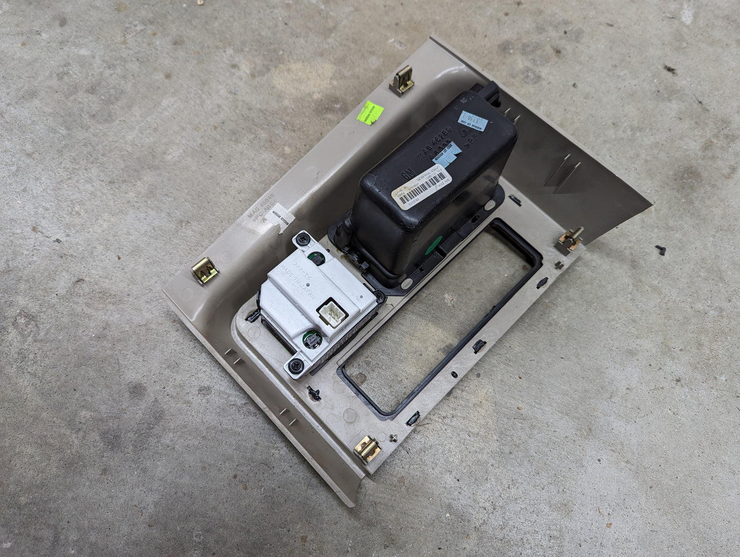 A General Motors OEM Center Console Front Bin Woodgrain Trim with Clock in Light Beige for 2003-2006 Cadillac Escalade lies on a concrete floor, featuring mounting slots and an opening for components.