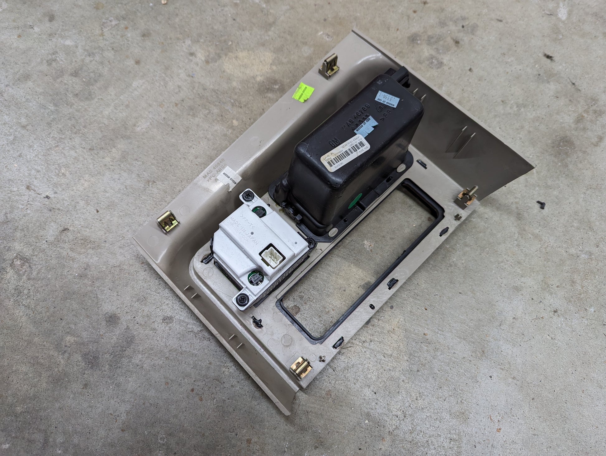 A General Motors OEM Center Console Front Bin Woodgrain Trim with Clock in Light Beige for 2003-2006 Cadillac Escalade lies on a concrete floor, featuring mounting slots and an opening for components.