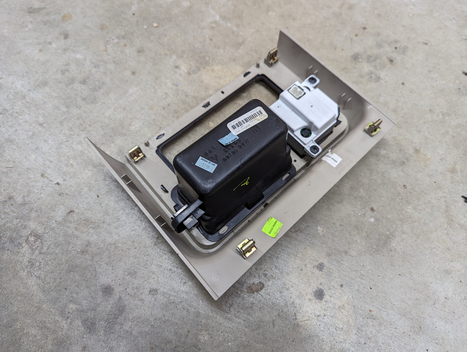 A General Motors OEM Center Console Front Bin Woodgrain Trim with Clock in Light Beige for 2003-2006 Cadillac Escalade rests on a concrete floor, featuring labels, a barcode, connectors, and metal clips.