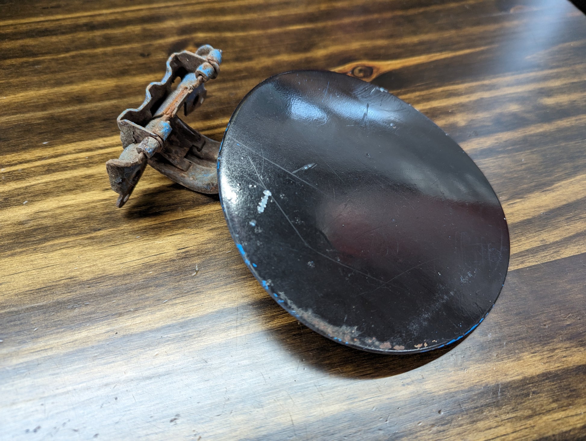 A rusty metal clamp is attached to a General Motors OEM Fuel Filler Neck Door Gas Tank Cover in Black for 2001-2004 Chevy S10 & GMC Sonoma, showing scratches and traces of blue paint, resting on a wooden surface.