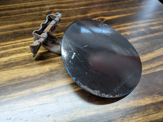 A rusty metal clamp is attached to a General Motors OEM Fuel Filler Neck Door Gas Tank Cover in Black for 2001-2004 Chevy S10 & GMC Sonoma, showing scratches and traces of blue paint, resting on a wooden surface.